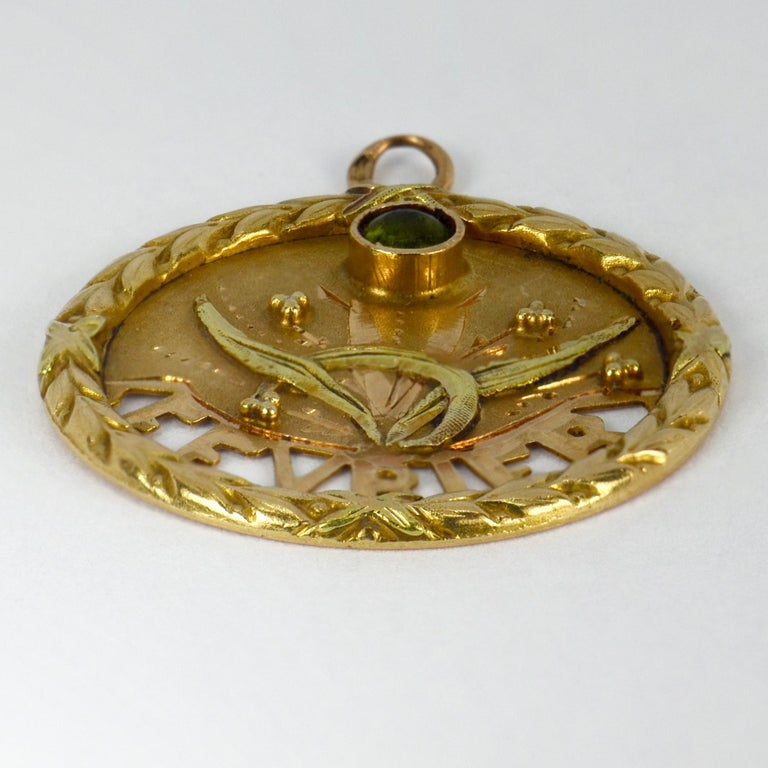 A French 18 karat (18K) gold charm pendant designed as a rose gold disc with a yellow gold laurel frame banded with ribbons, with the word ‘Fevrier’ (February) pierced out. Beneath a collet set cabochon green paste an iris (the flower of February)