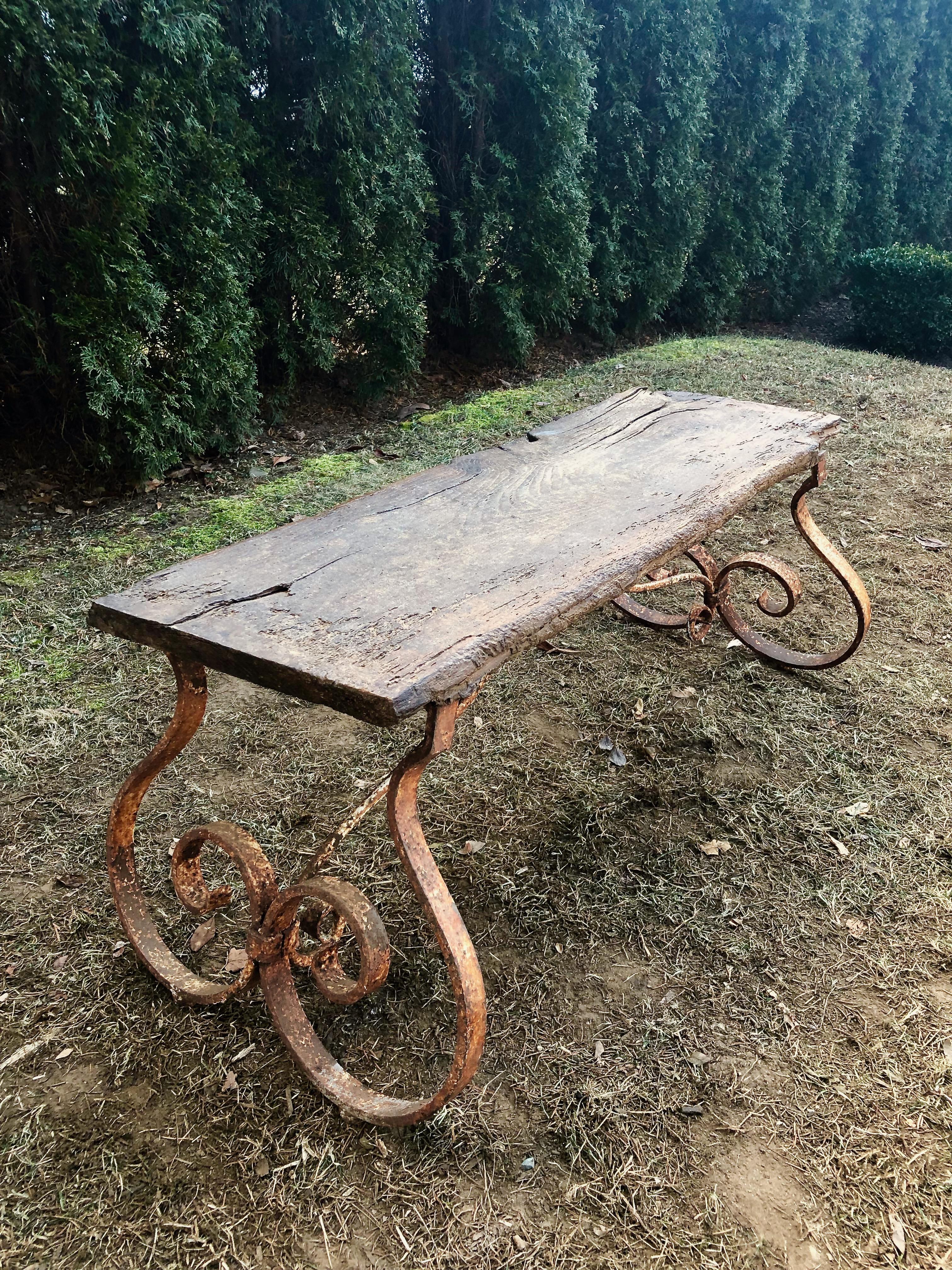 If you are looking for rustic, this piece is sure to please! We believe the wooden slab may be chestnut, and the legs are thick, hand forged wrought iron that have been attached to the underside of the top with braces. The wood sports a wonderful