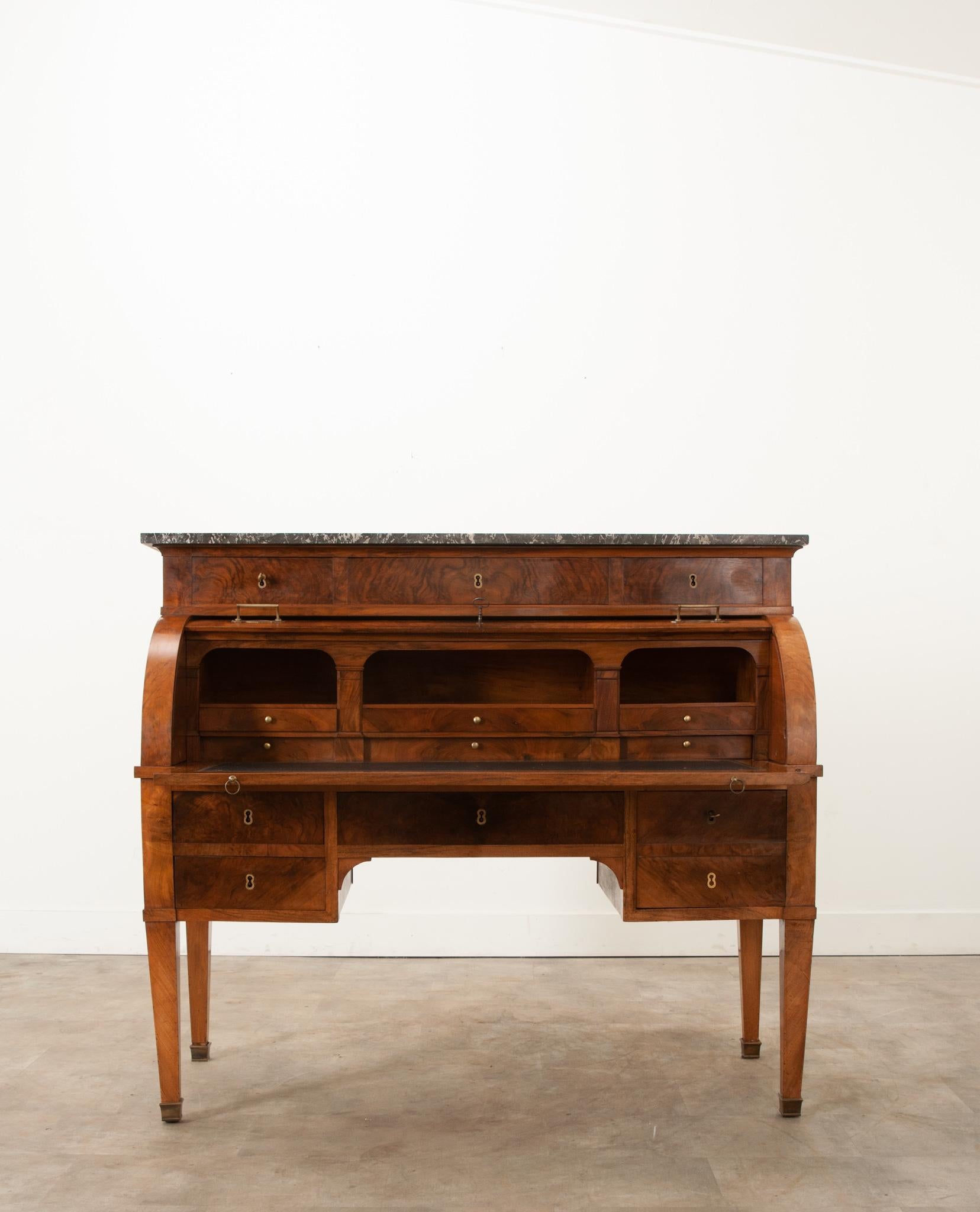 A handsome and classically designed French Directoire “D” roll top desk. The desk is topped with a polished piece of  St. Anne gris marble. Directly beneath the marble are three drawers with beautiful bookmatched burl mahogany drawer fronts and