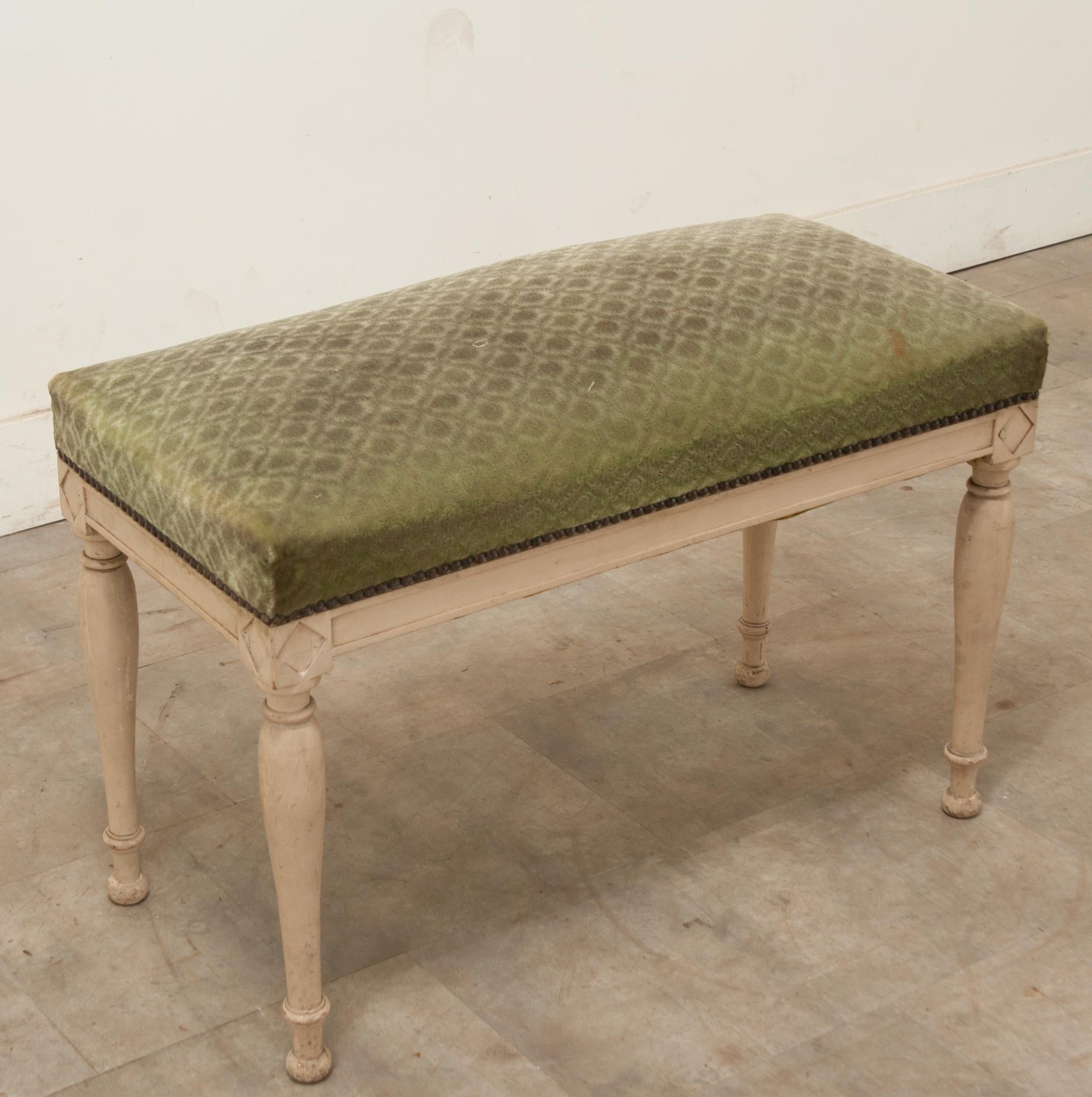 A French Directoire period painted bench made during the late 1700’s. This bench is upholstered in a worn green velvet upholstery with a nailhead trim. The frame has diamond carvings on the corners above its turned legs connected to turned ball