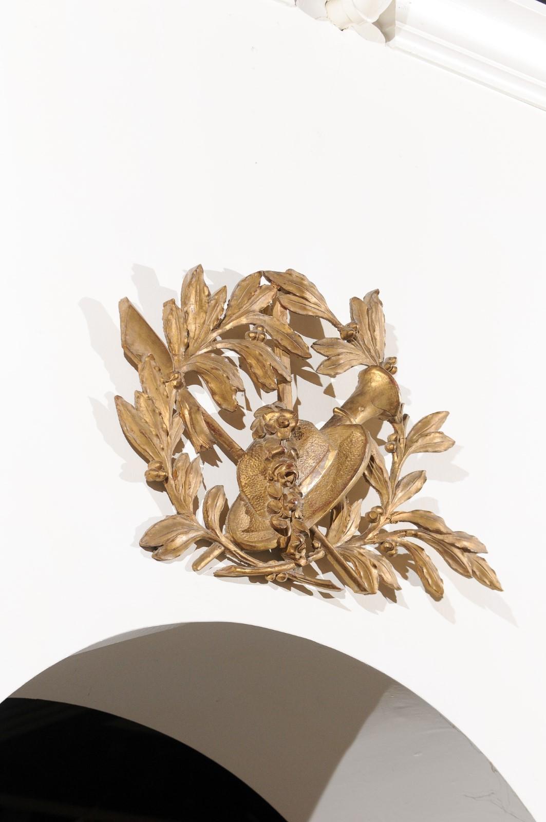 A French hand carved giltwood wall decoration from the 18th century depicting a straw hat, olive branches and instruments. Charming our eyes with its golden tone and exquisitely carved ornaments, this French 18th century wall decoration is centered