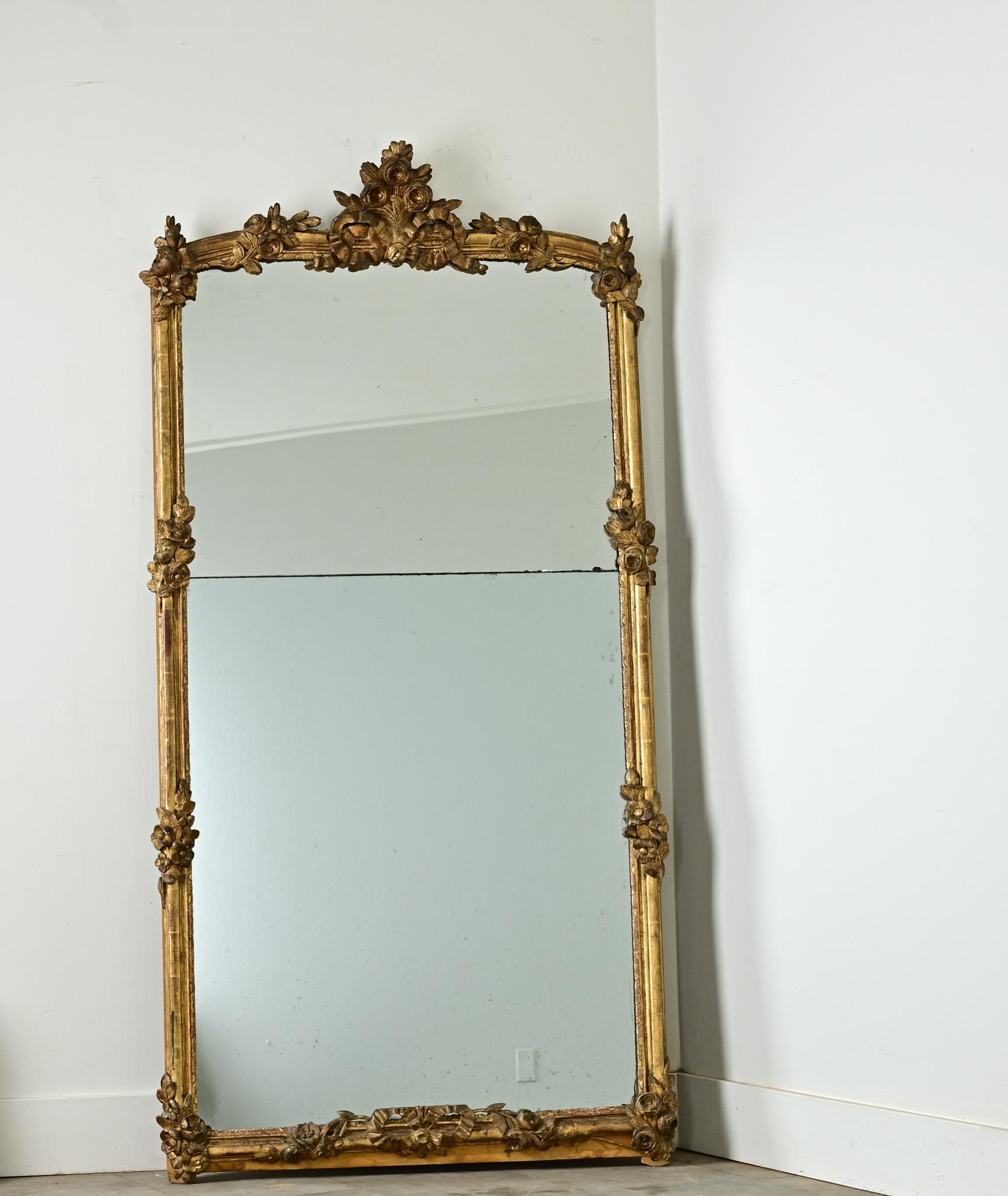 Décadent et romantique, ce miroir Louis XV est animé par des sculptures florales arrondies et luxuriantes qui se détachent du cadre doré par des rubans. Deux plaques de miroir présentent des rousseurs, une oxydation qui correspond aux cavités