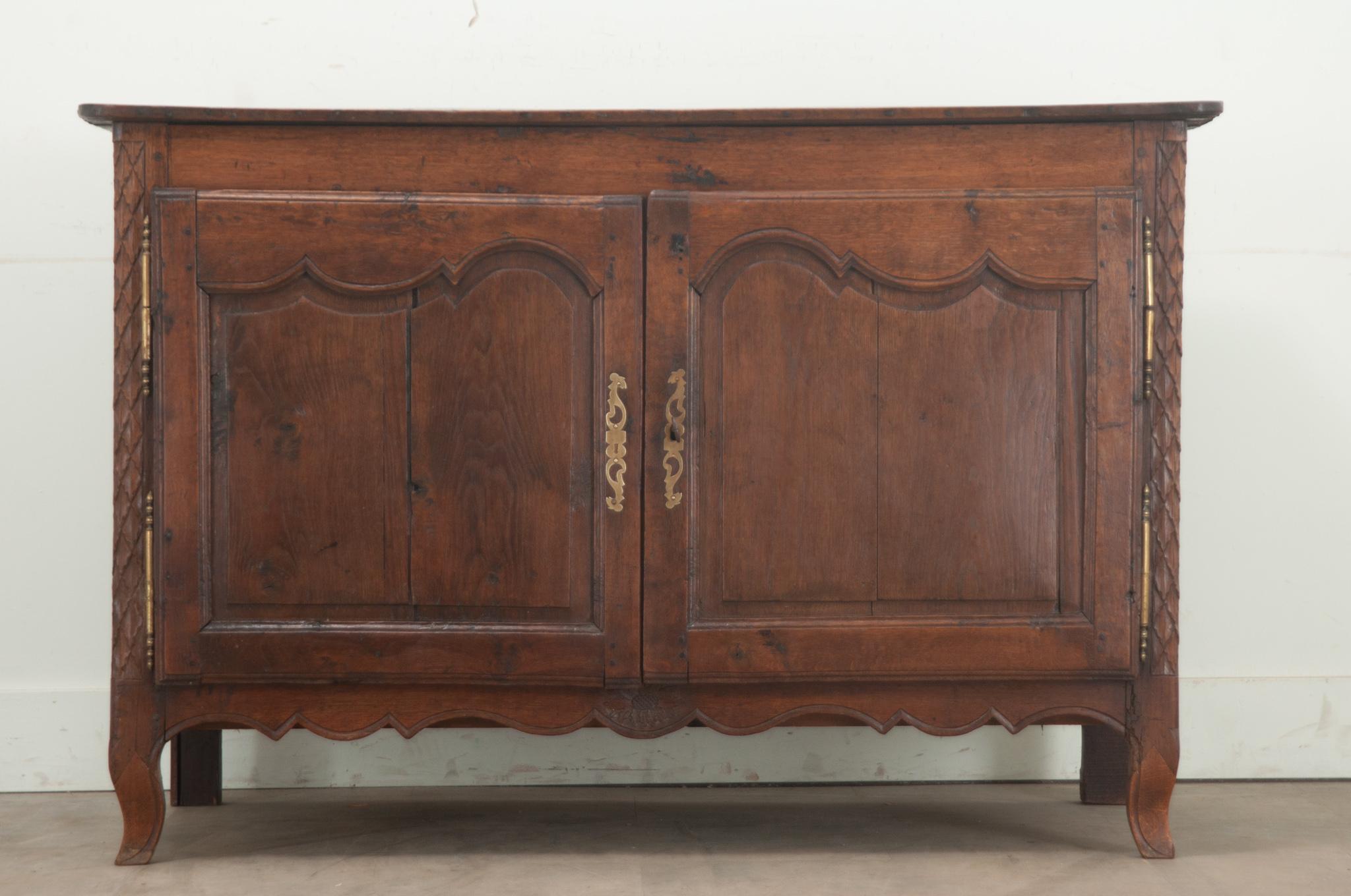 A French Louis XV period solid oak buffet. This buffet has shapely carved doors over a scalloped apron with brass barrel hinges and hand cut escutcheon plates. The doors open with a working lock and key to reveal one fixed shelf at 19 ½” D. Cleaned