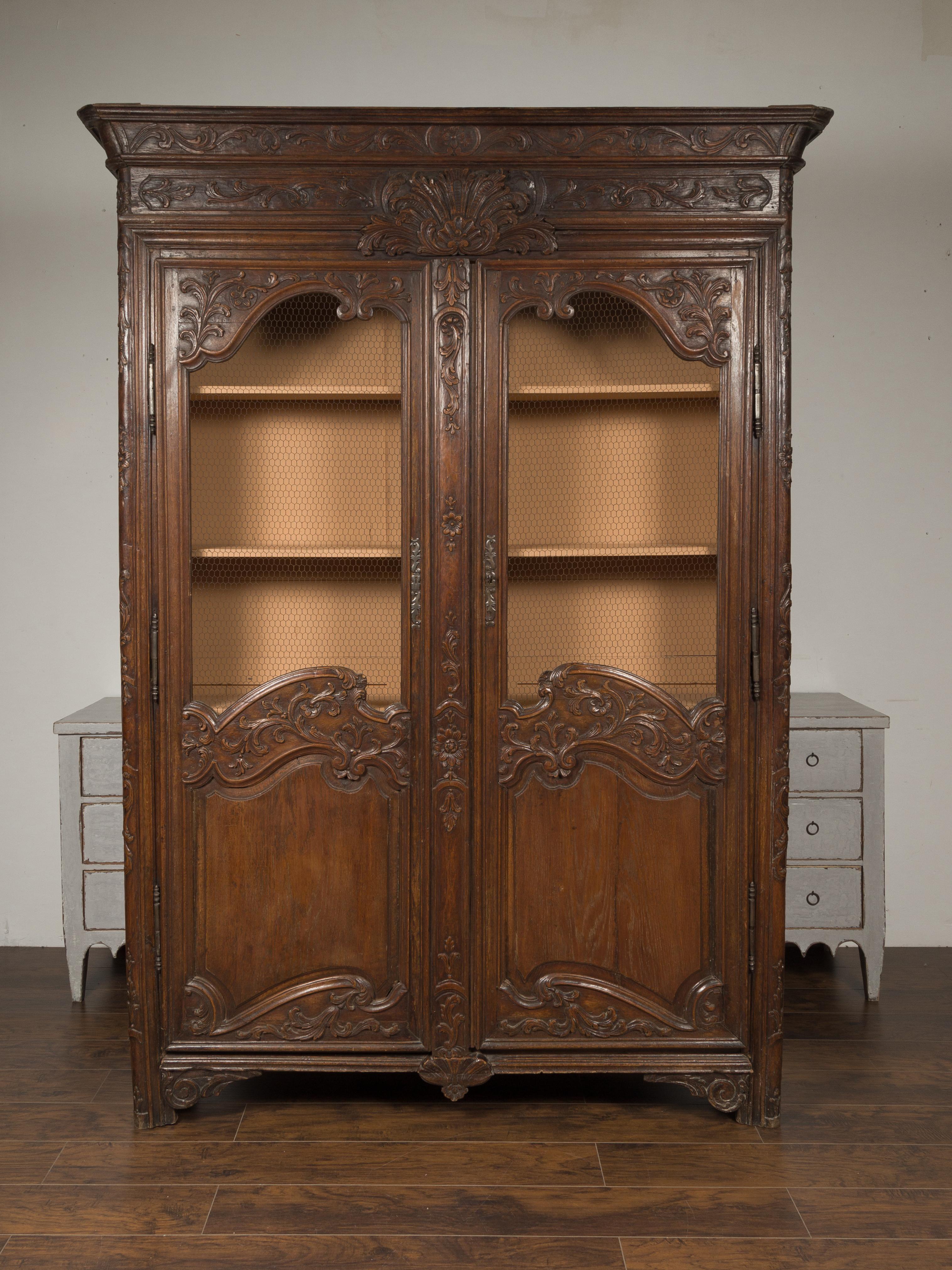 This French 18th century Louis XV narrow wooden bibliothèque (bookcase in French) features two carved doors with chicken wire below a crown molding adorned with a rinceaux frieze and a central carved motif made of a bouquet of acanthus leaves and