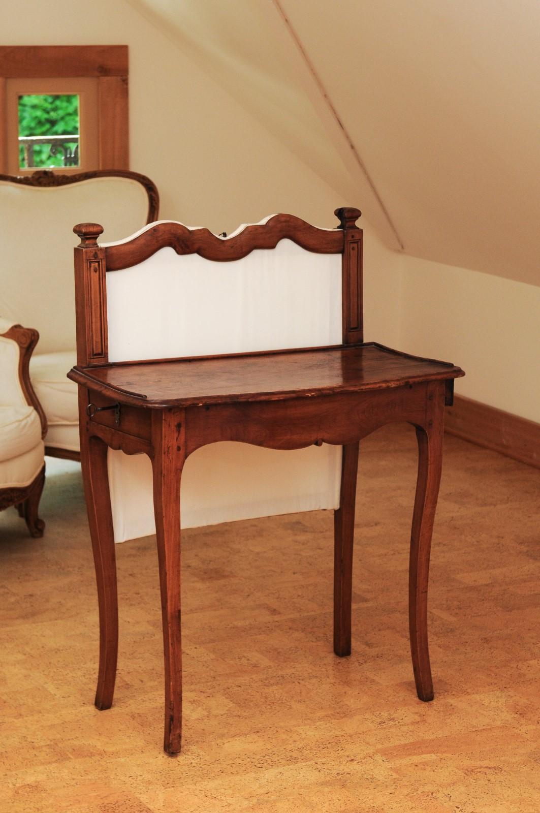 A French Louis XV period walnut par-feu writing table from the 18th century, with petite finials, side drawers and curving legs. Created in France during the reign of King Louis XV, this walnut par-feu desk features an upholstered back topped with