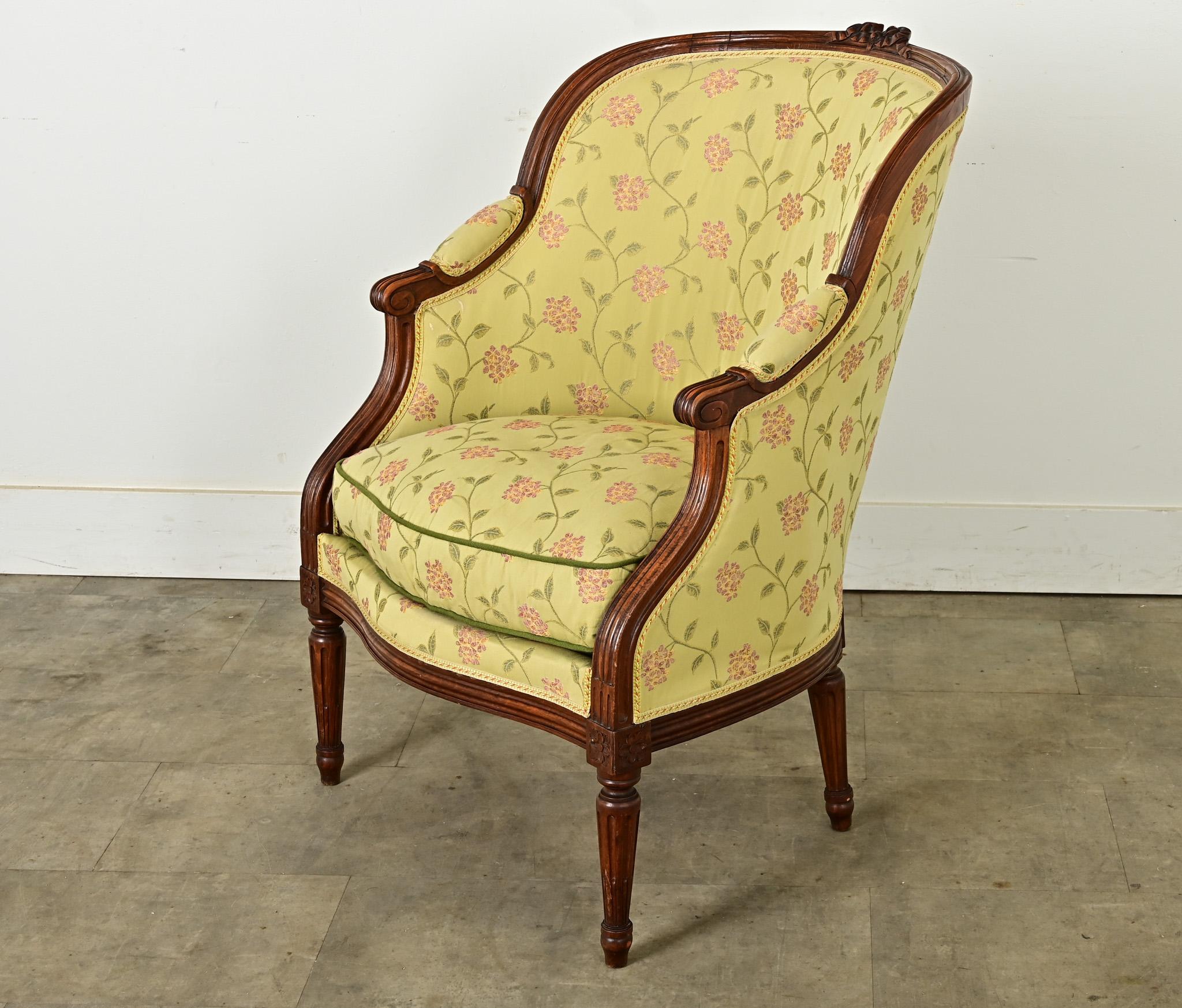 A delightful French Louis XVI-style bergere from circa 1780 in floral upholstery of wispy pink and gold blooms on pistachio background, outlined by the dark mahogany carved frame. Crowned with a bow-shaped carving atop the barrel back, fluted