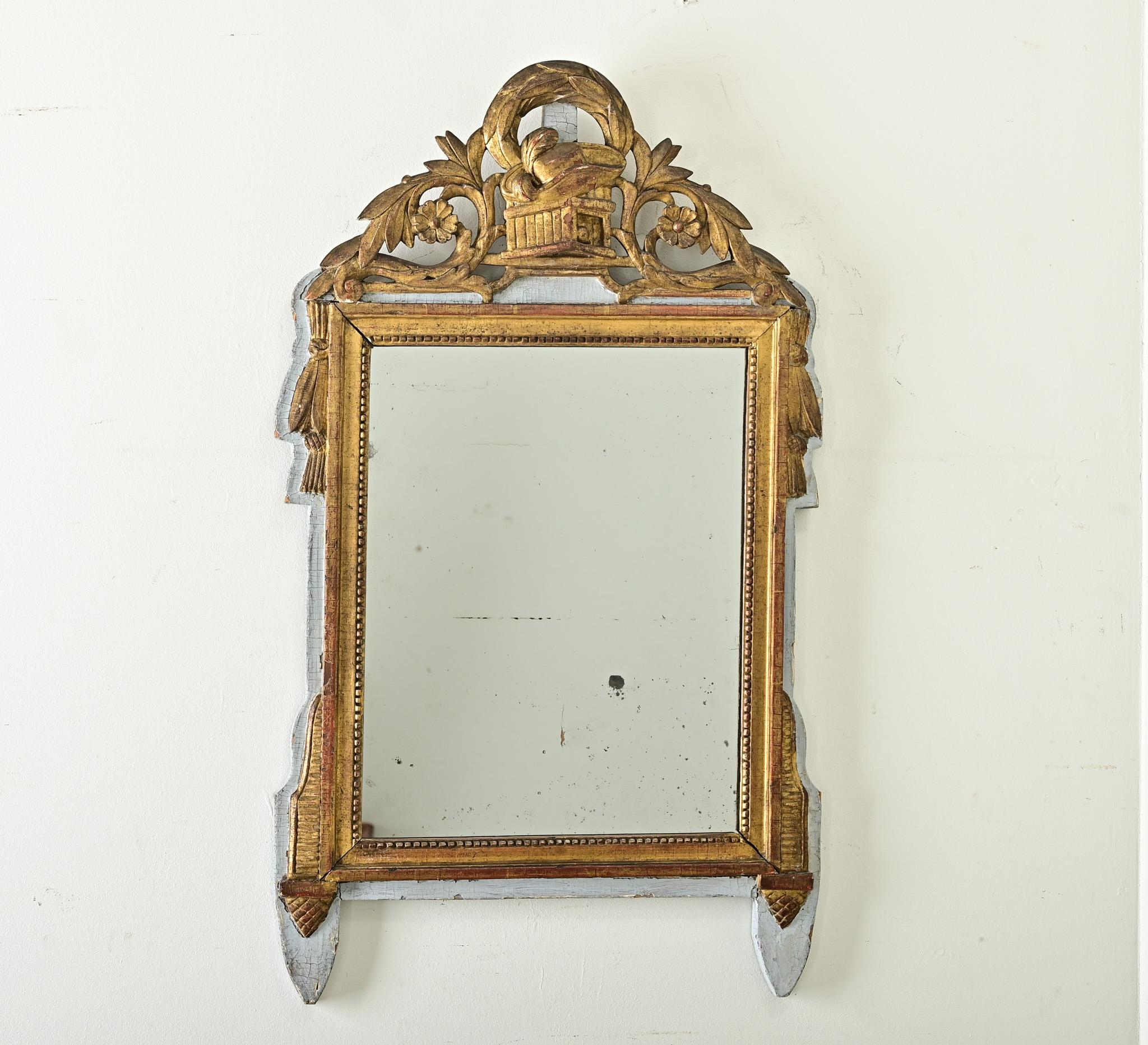An eyecatching French trumeau style mirror, Louis XVI period (18th century). The painted and giltwood frame has an ornate crest at the top with seemingly disparate imagery such as a hat with ostrich feathers, a laurel wreath, and flowering vines