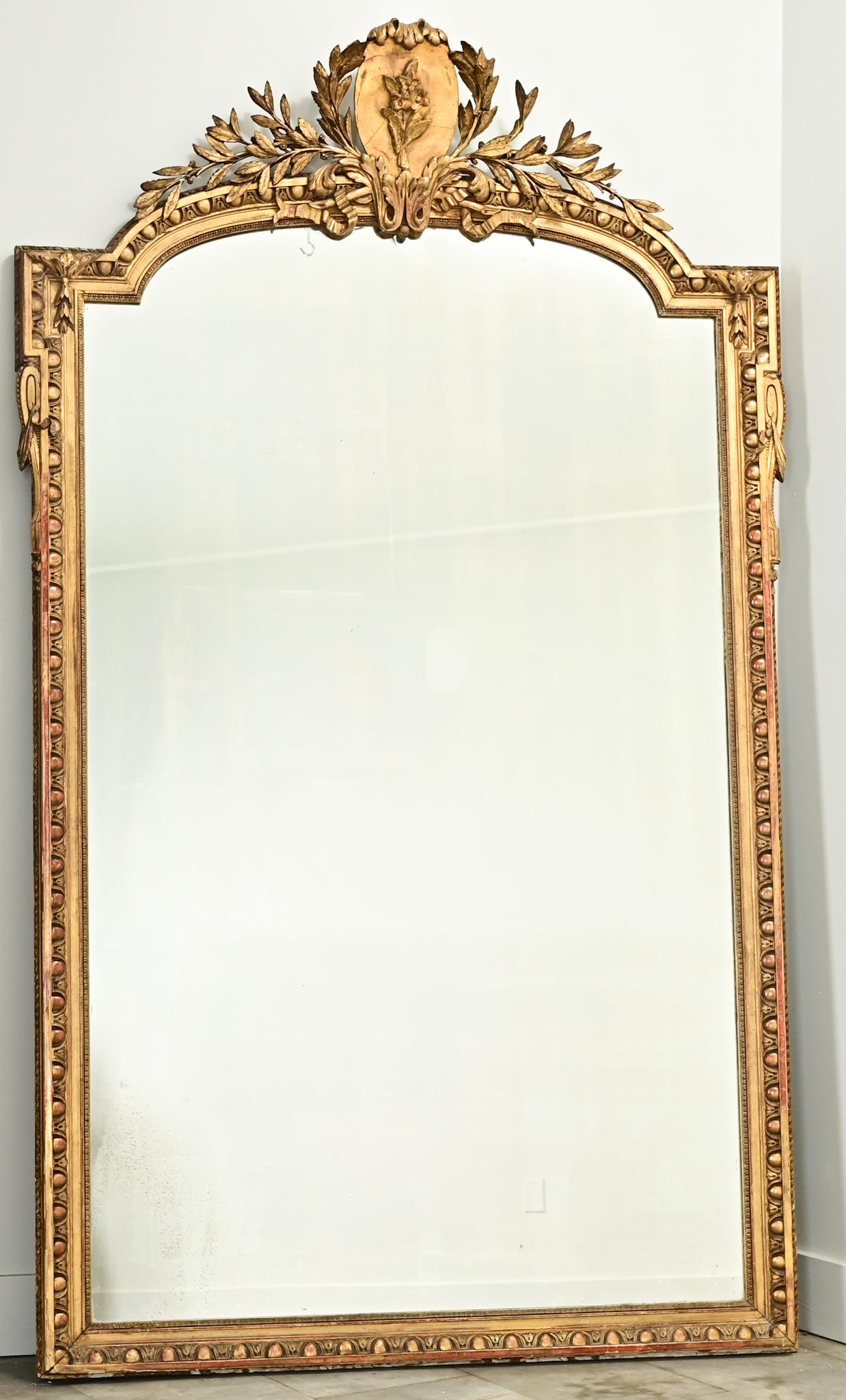 A large French Louis XVI gold gilt mirror made in 18th Century France. The mirror has a carved plaster crest with a center floral cartouche and laurel leaf branches. The center top of the frame is rounded with square corners and embellished with egg