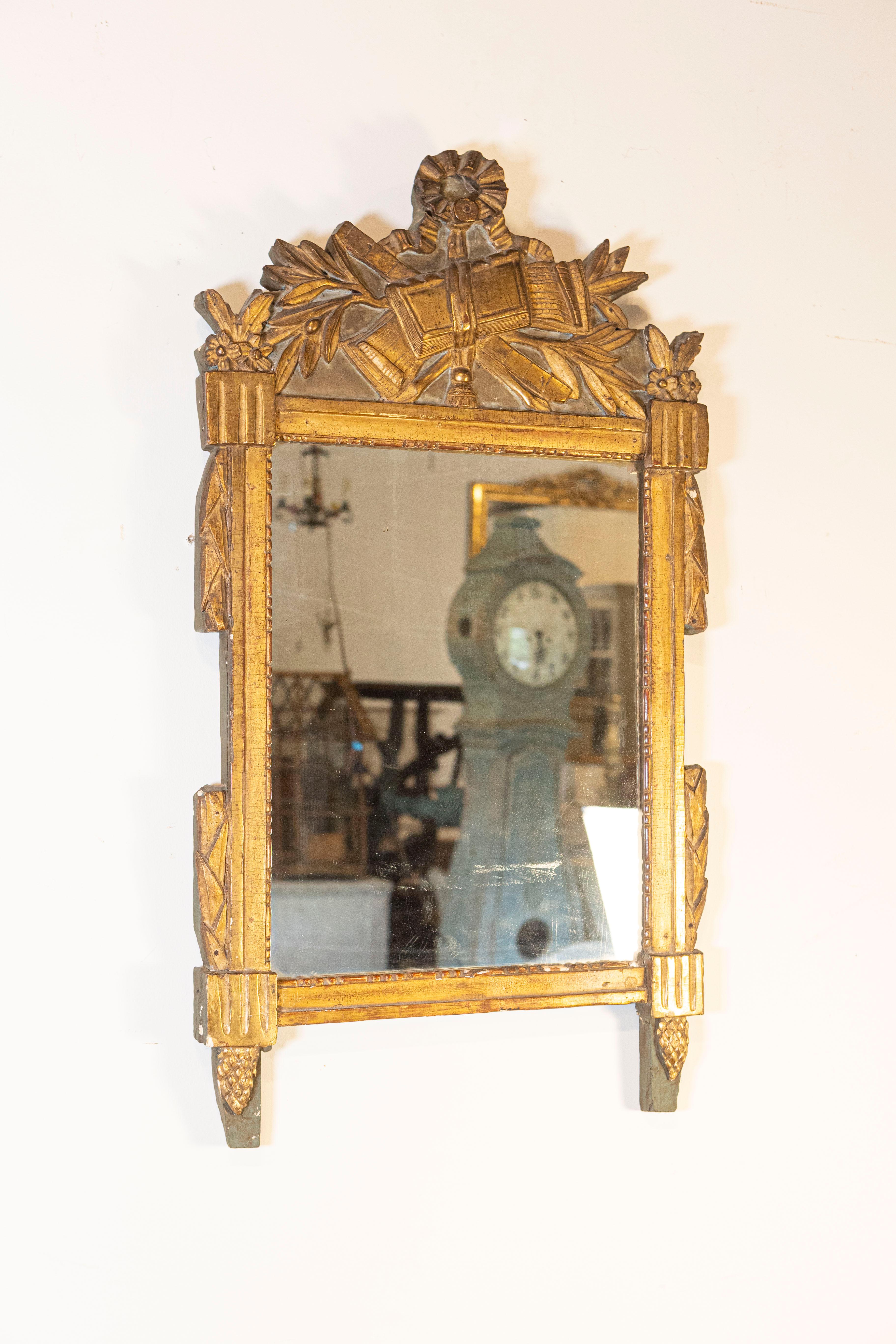 Miroir en bois doré d'époque Louis XVI vers 1790 avec allégorie des arts libéraux sculptée en ruban, motifs de feuilles de laurier et de pommes de pin, fond peint, miroir légèrement vieilli et caractère rustique. Voici un magnifique miroir en bois