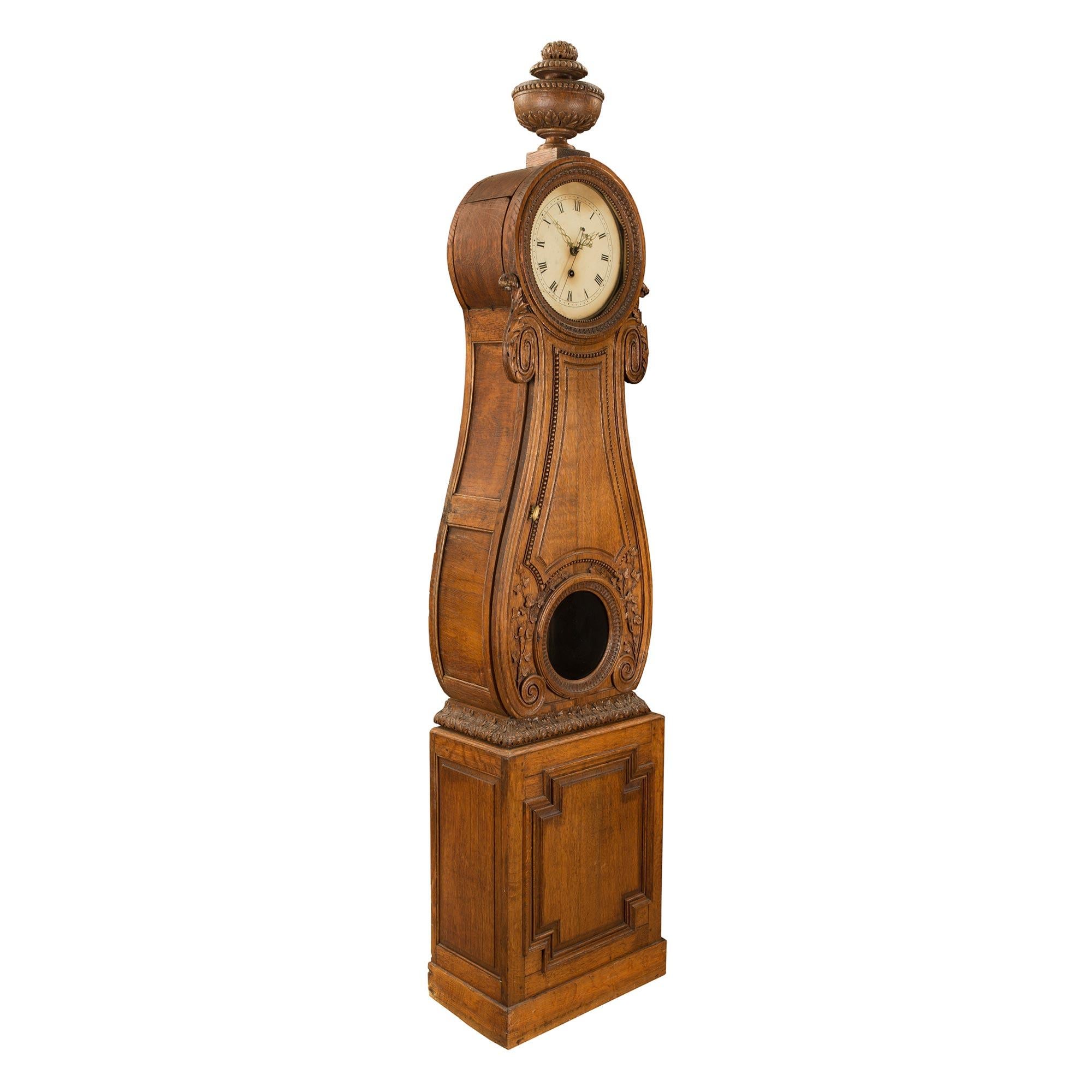 A handsome French 18th century Louis XVI period oak grandfather clock. The clock is raised by a square mottled base with an elegant carved recessed design. At the center is a finely carved acanthus leaf border below the circular glass display with