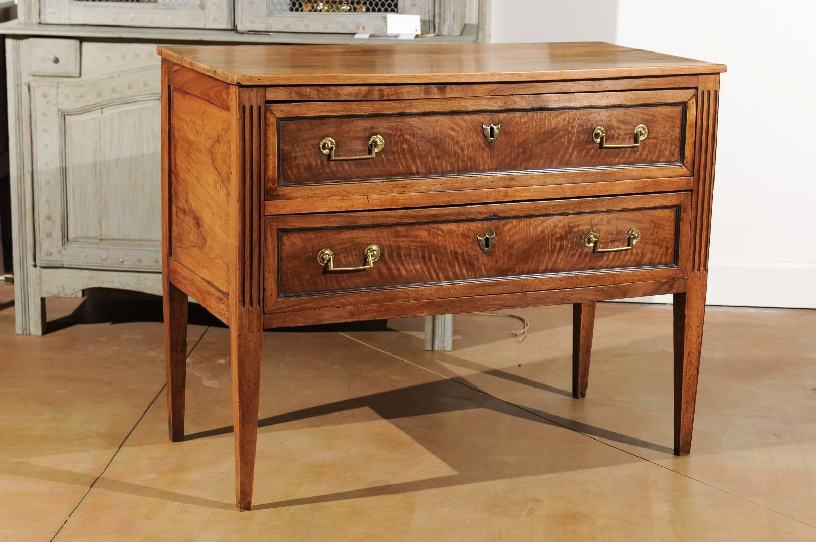 A French Louis XVI period two-drawer walnut commode from the third quarter of the 18th century, with fluted side posts and tapered legs. This French walnut commode features a rectangular planked top sitting above two dovetailed drawers. Each drawer