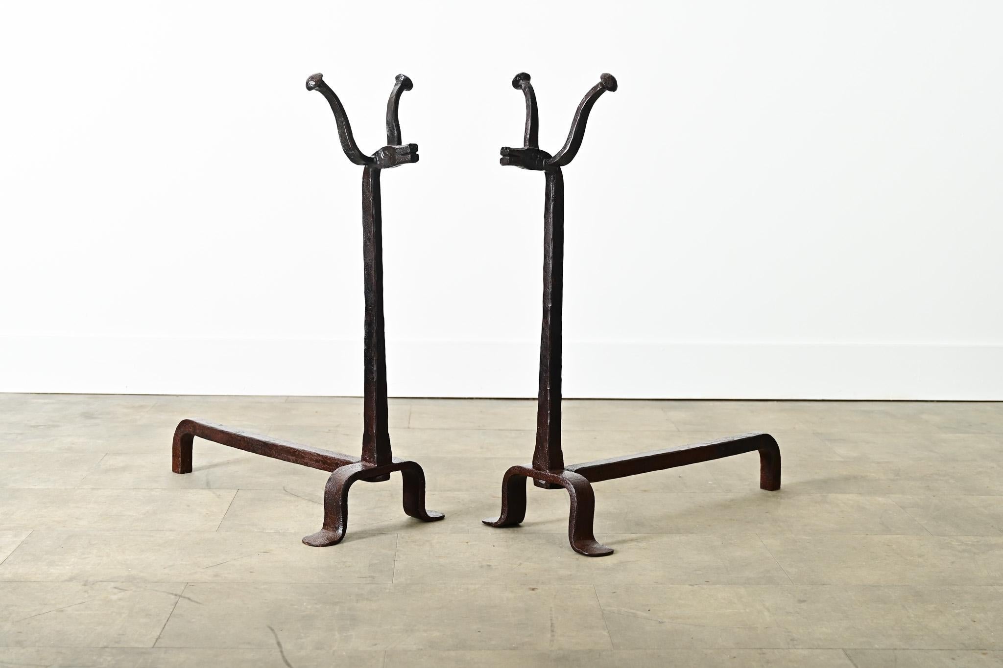 A very unique pair of 18th century French wrought iron chenets–or andirons–forged with zoomorphic heads of bulls atop the otherwise square & straight proportions. The stylized animals feature a pair of horns mounted on their faces, complete with