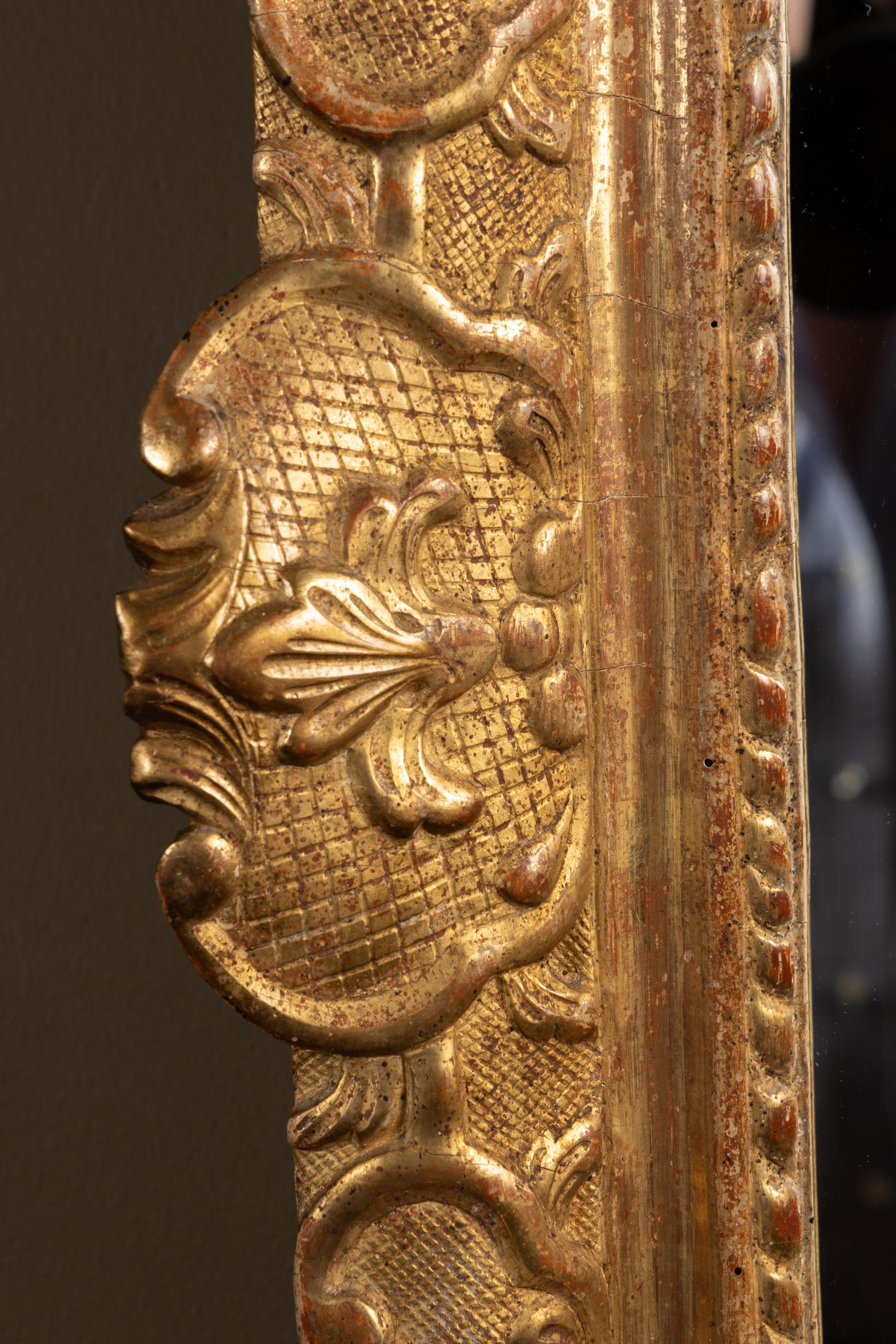 Miroir français du XVIIIe siècle de style Régence, sculpté à la feuille d'or, orné d'un motif héraldique au centre de la partie supérieure réticulée consistant en une floraison de drapeaux et de cornes, superposée à un grand casque, flanqué d'un