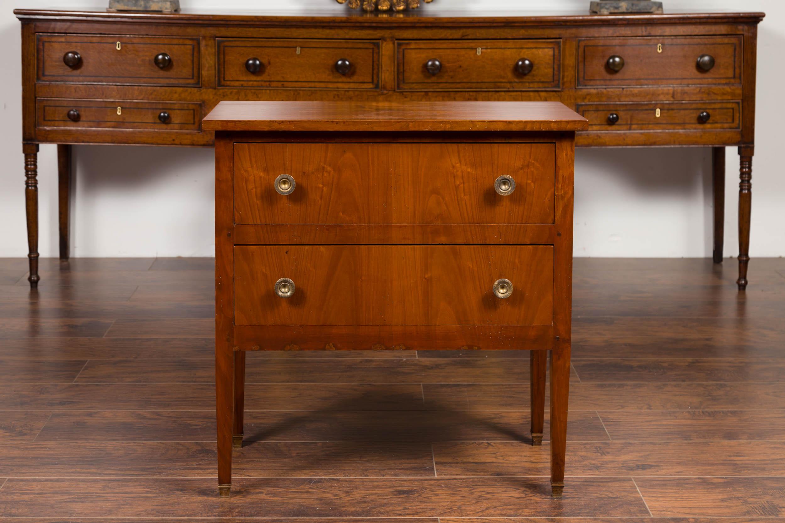 A French bookmark veneer two-drawer commode from the early 20th century, with petite brass feet. Born in France at the turn of the century, this wooden commode features a rectangular planked top overhanging two dovetailed drawers, each fitted with