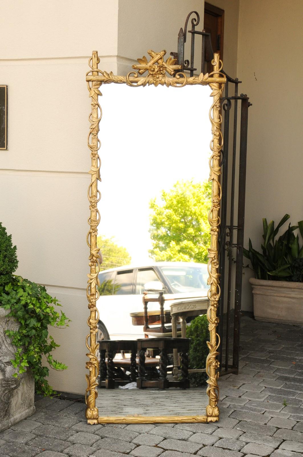 A French carved giltwood mirror from the early 20th century with faux branches and floral decor. Born in France during the early years of the 20th century, this exquisite mirror features a slender Silhouette, beautifully adorned with a giltwood