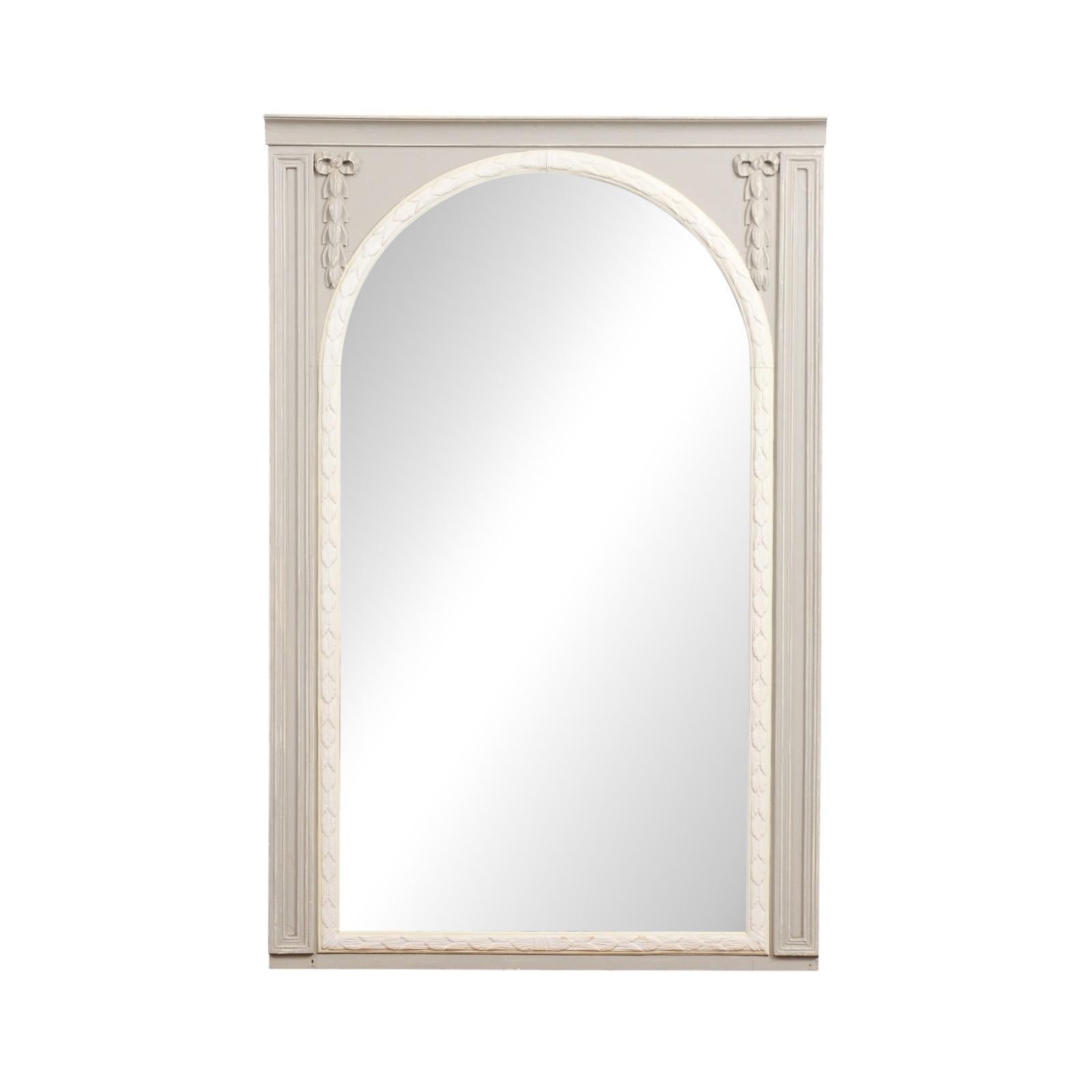 A French two-toned painted wood trumeau mirror from the early 20th century with carved foliage, ribbon-tied motifs and arched mirror plate. Created in France at the Turn of the Century, this trumeau mirror features a two-toned finish showcasing a