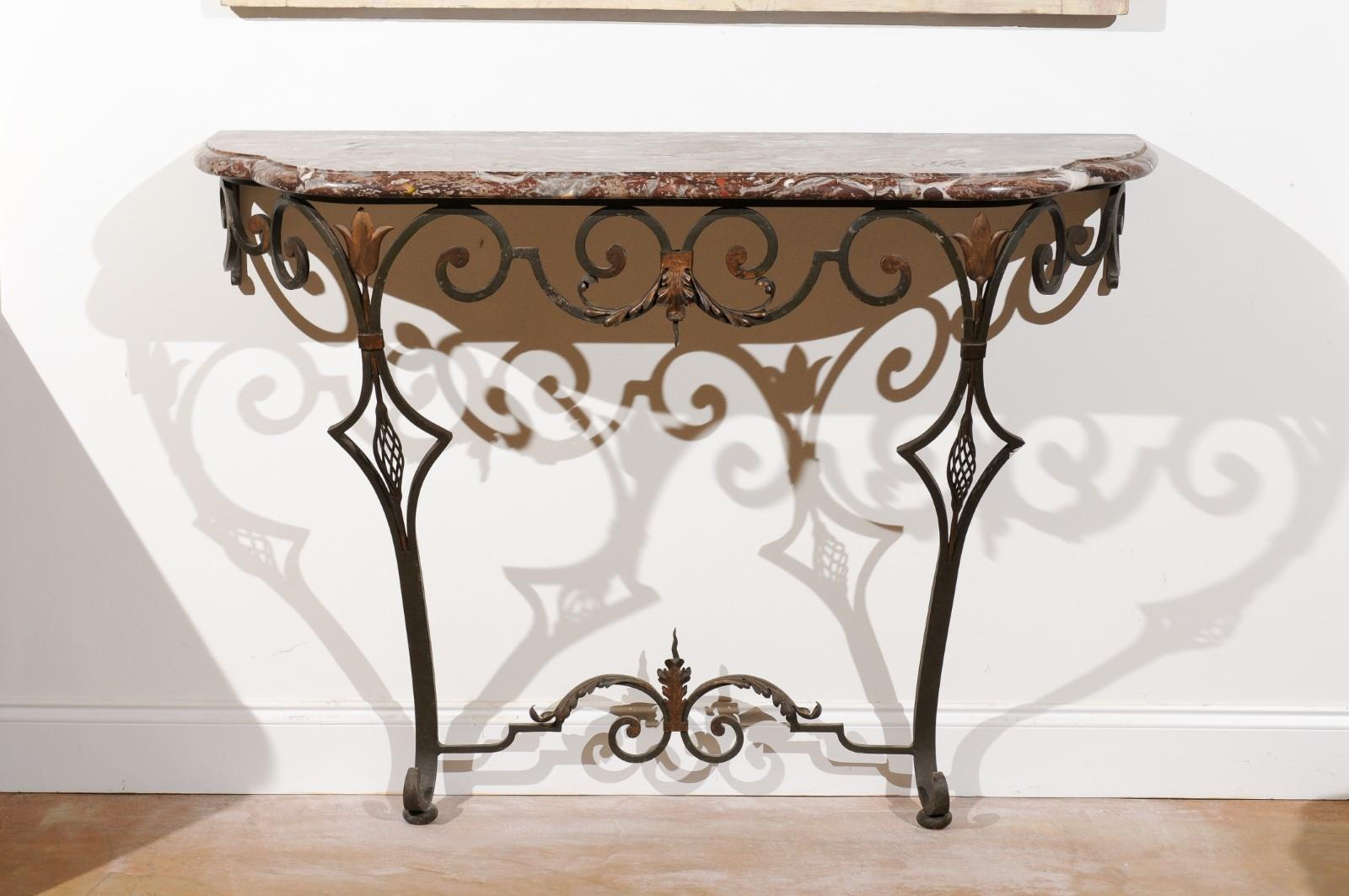 A French wrought iron console table from the early 20th century with variegated marble top and scrolling base. This exquisite French console table features a variegated red marble top with rounded edges, sitting above a graceful wrought iron