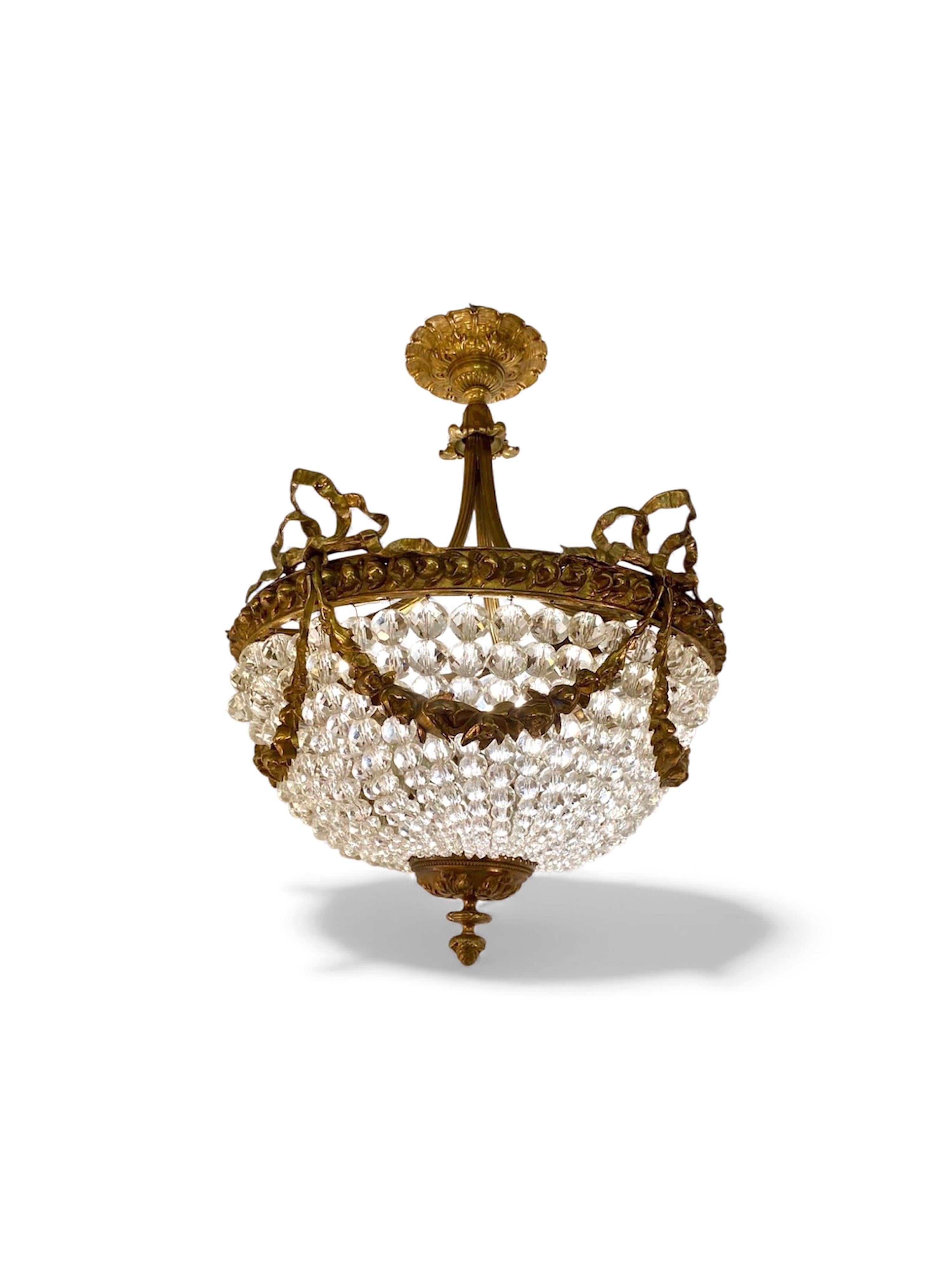 Ce lustre français du début du 19e siècle, de style Empire, présente un motif de panier en cristal, des guirlandes, des couronnes de laurier et des nœuds en métal. Les trois bras  Les bras relient le corps à la coupelle du plafond. 
Tout cela vous