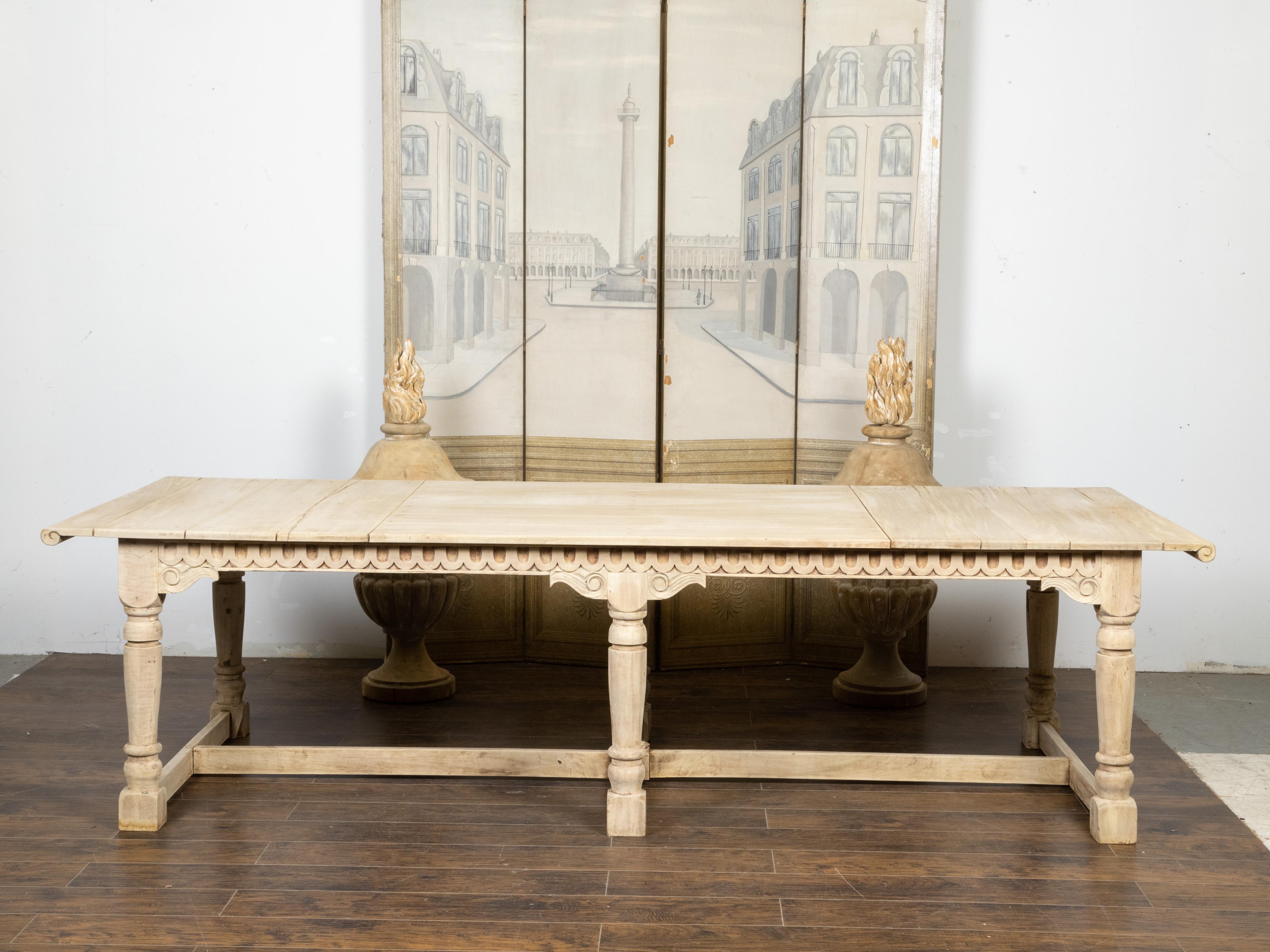A French bleached walnut dining room table from the early 20th century, with carved apron, scoop motifs, baluster legs, scrolling accents and double H-Form cross stretcher. Created in France during the first quarter of the 20th century, this walnut