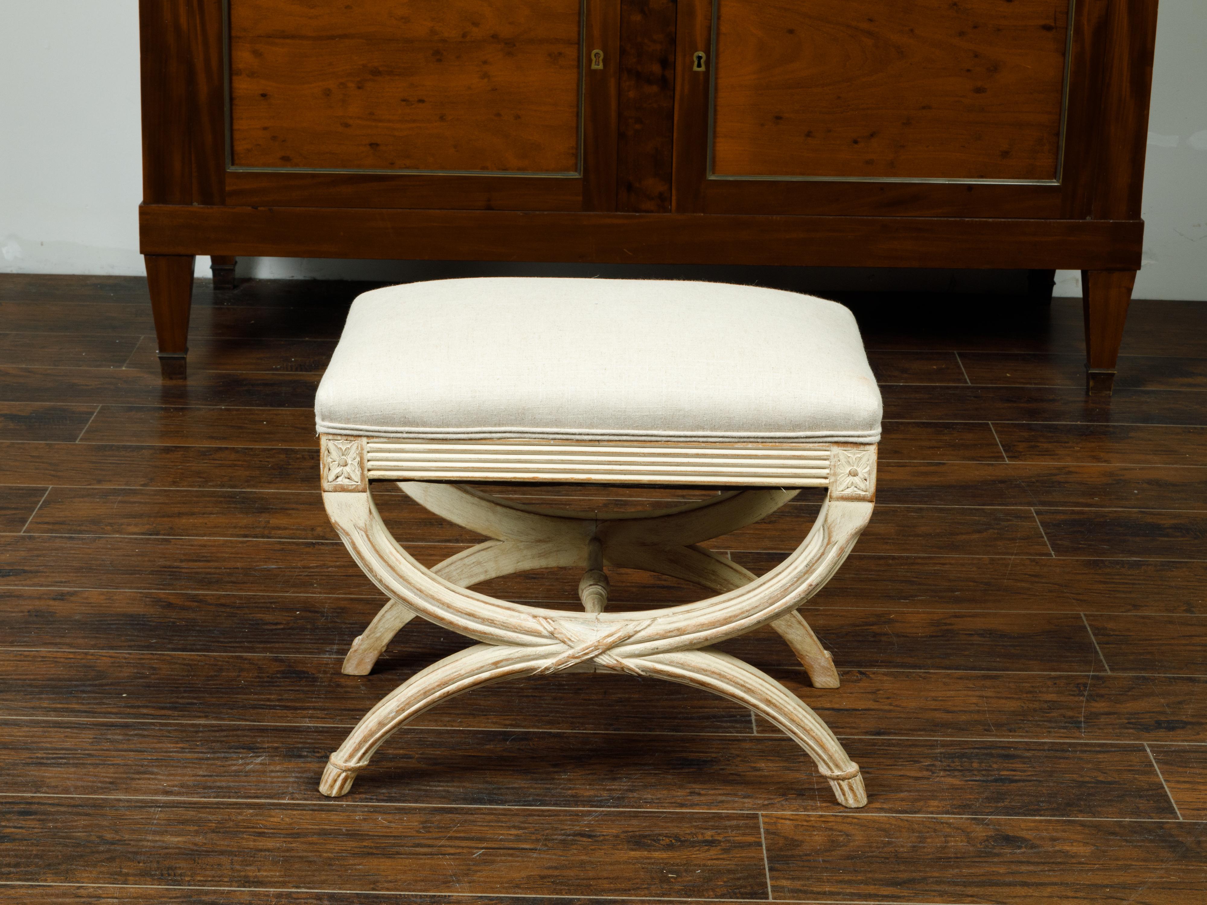 A French Empire style painted wood curule stool from the early 20th century, with carved rosettes and new upholstery. Created in France during the first quarter of the 20th century, this painted stool features a rectangular seat newly recovered with