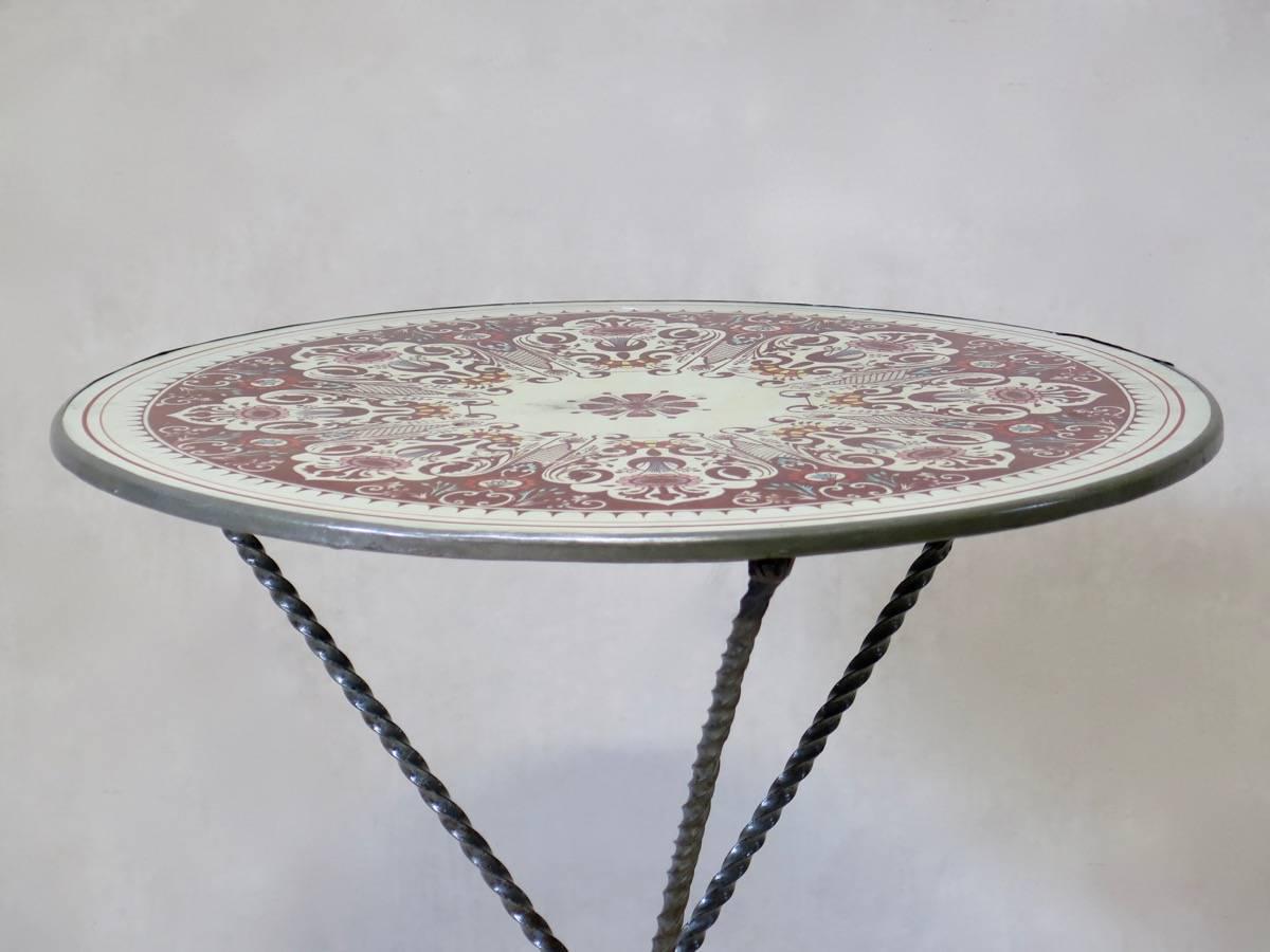 Petite table ronde tripode en guéridon, avec un plateau en métal émaillé à motifs et des pieds en fer torsadé. La table était auparavant pliable.