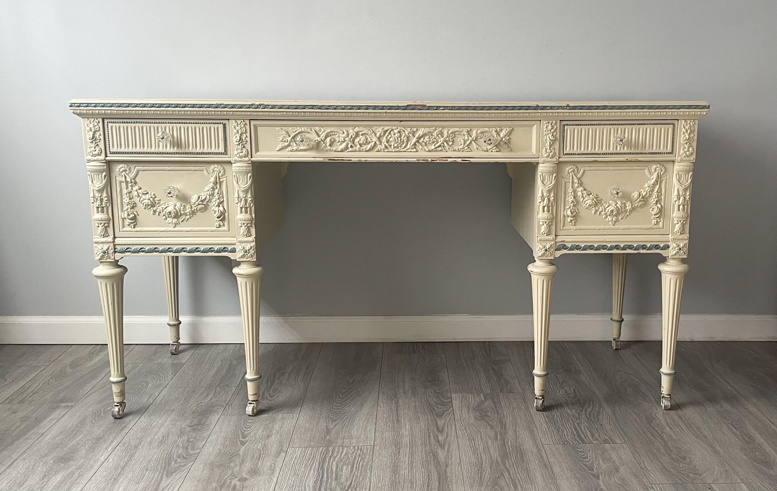 Beautiful, French 1920s painted vanity desk in the Louis XVI style.

The desk features intricately carved classic Louis XVI stylings and an ivory with blue trim paint finish. The glass knobs on the five drawers were a later addition but compliment