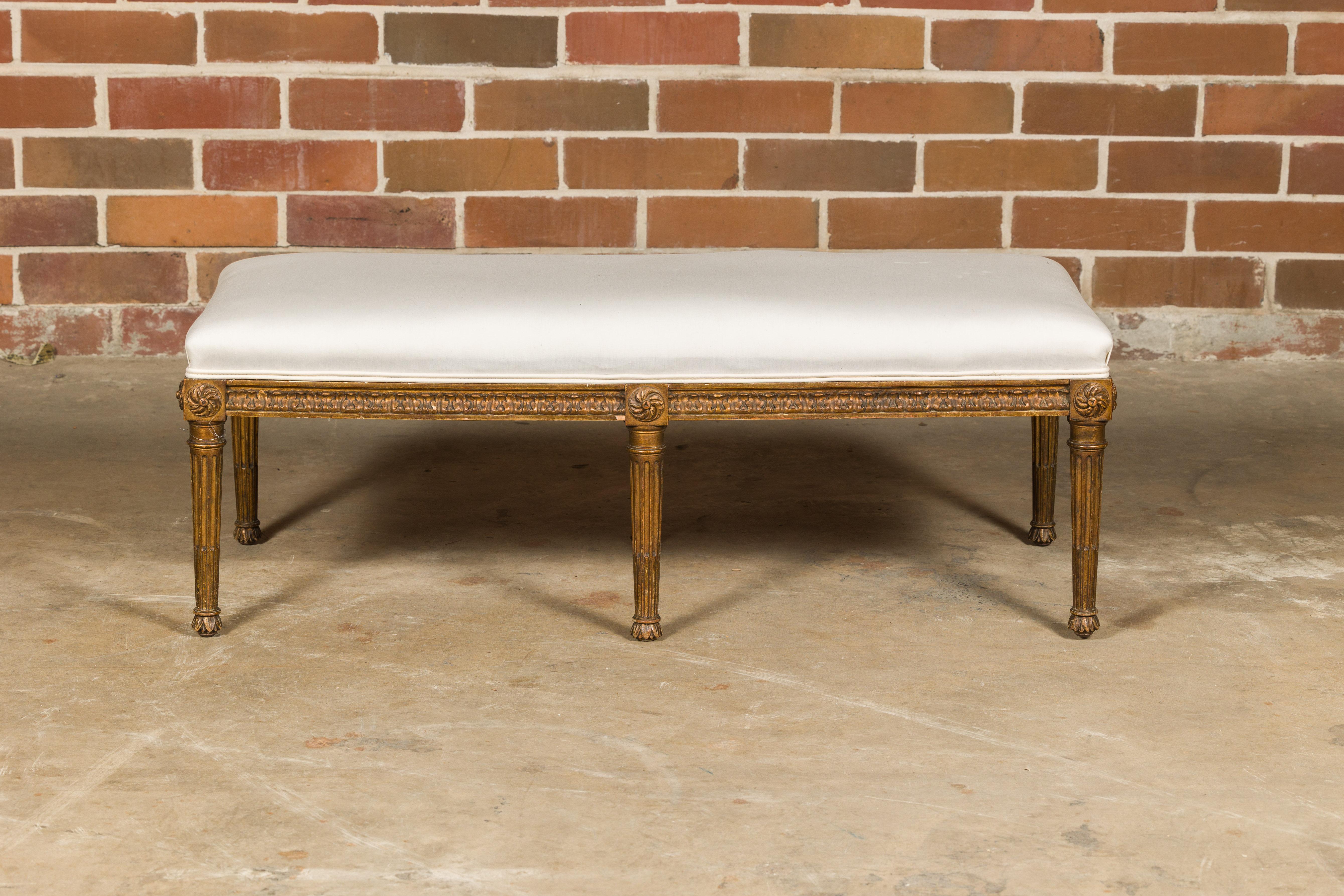 A French Neoclassical style gilded wood bench from circa 1920 with carved frieze, rosettes, fluted legs and custom muslin upholstery. This French Neoclassical style gilded wood bench, crafted around 1920, is a timeless testament to the elegance and