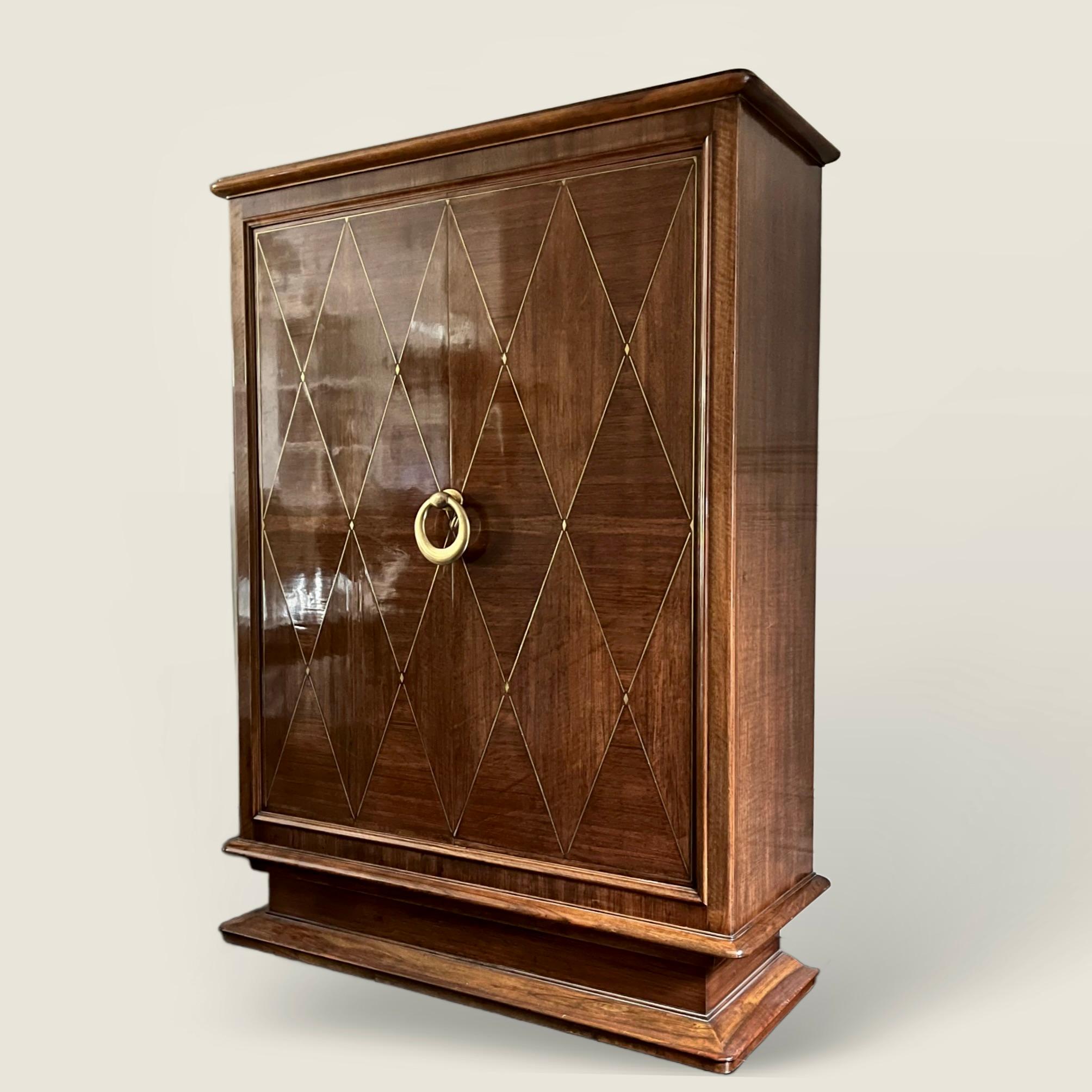 Armadietto francese degli anni '40 intarsiato con un raffinato motivo a rombi in ottone, rifinito con una grande e pesante maniglia circolare in ottone in posizione centrale, con serratura e chiave originali. 
L'interno è dotato di due appendiabiti