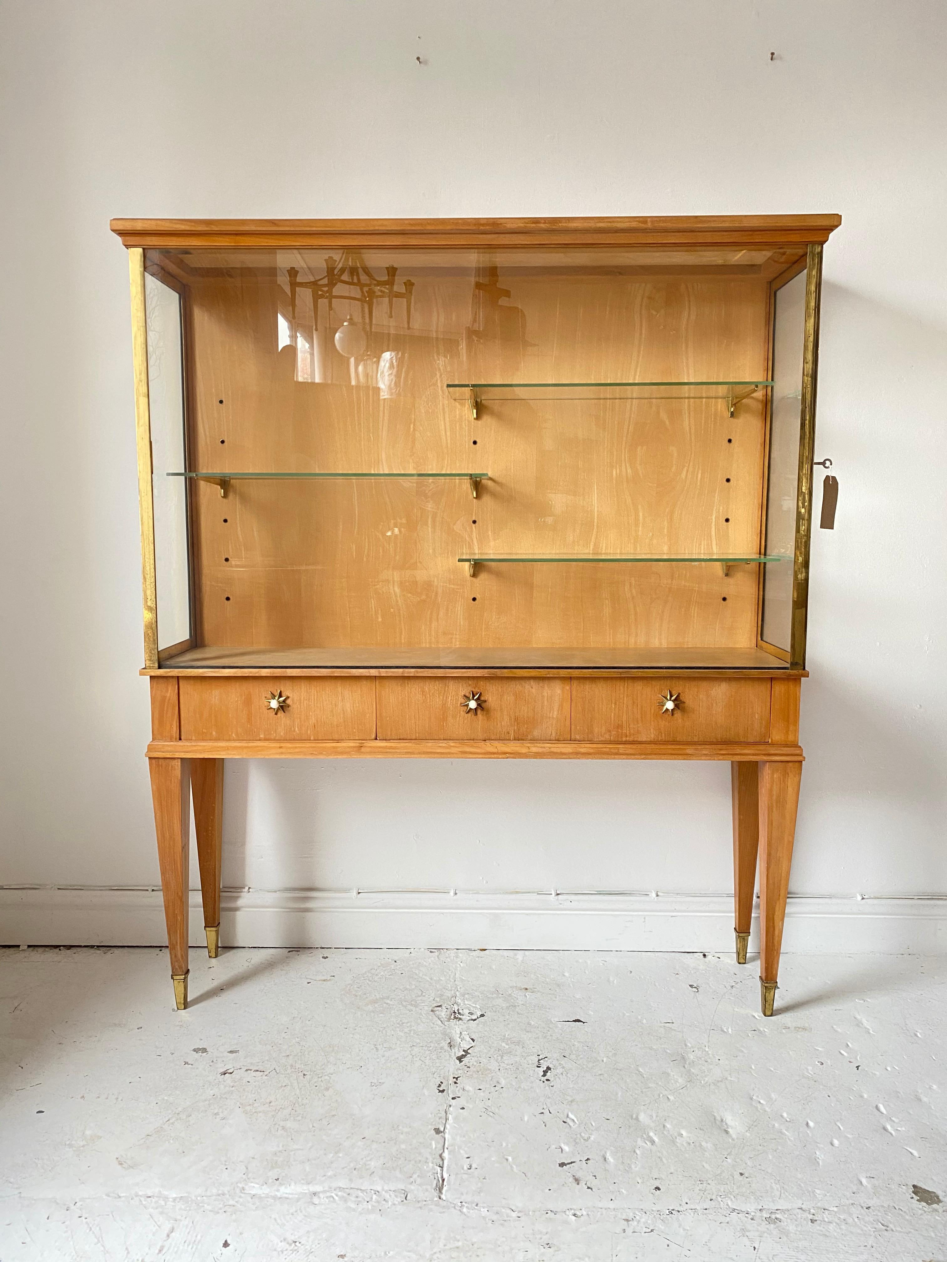 French 1940s brass trimmed vitrine cabinet, side opening with keys. Adjustable glass shelves with brass supports. The drawers open with striking star pulls. The feet are capped with brass. 

 This gorgeous cabinet has shades of the classic French