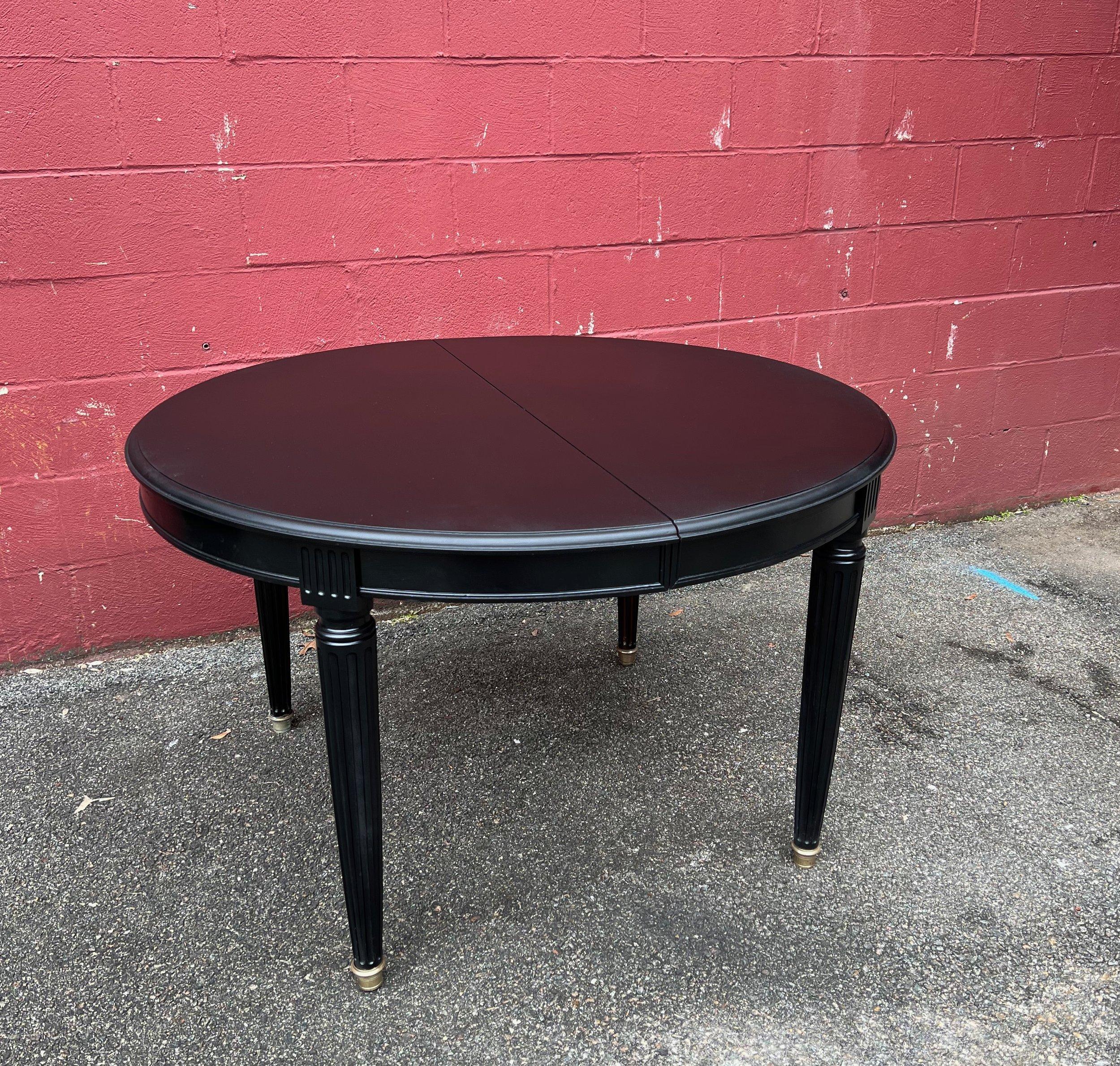 This elegant circular French 1940s neoclassical style mahogany veneered dining table features graceful tapered legs on brass sabots and it is has been recently refinished in a rich deep ebony. The table is equipped with stretchers that allow for two