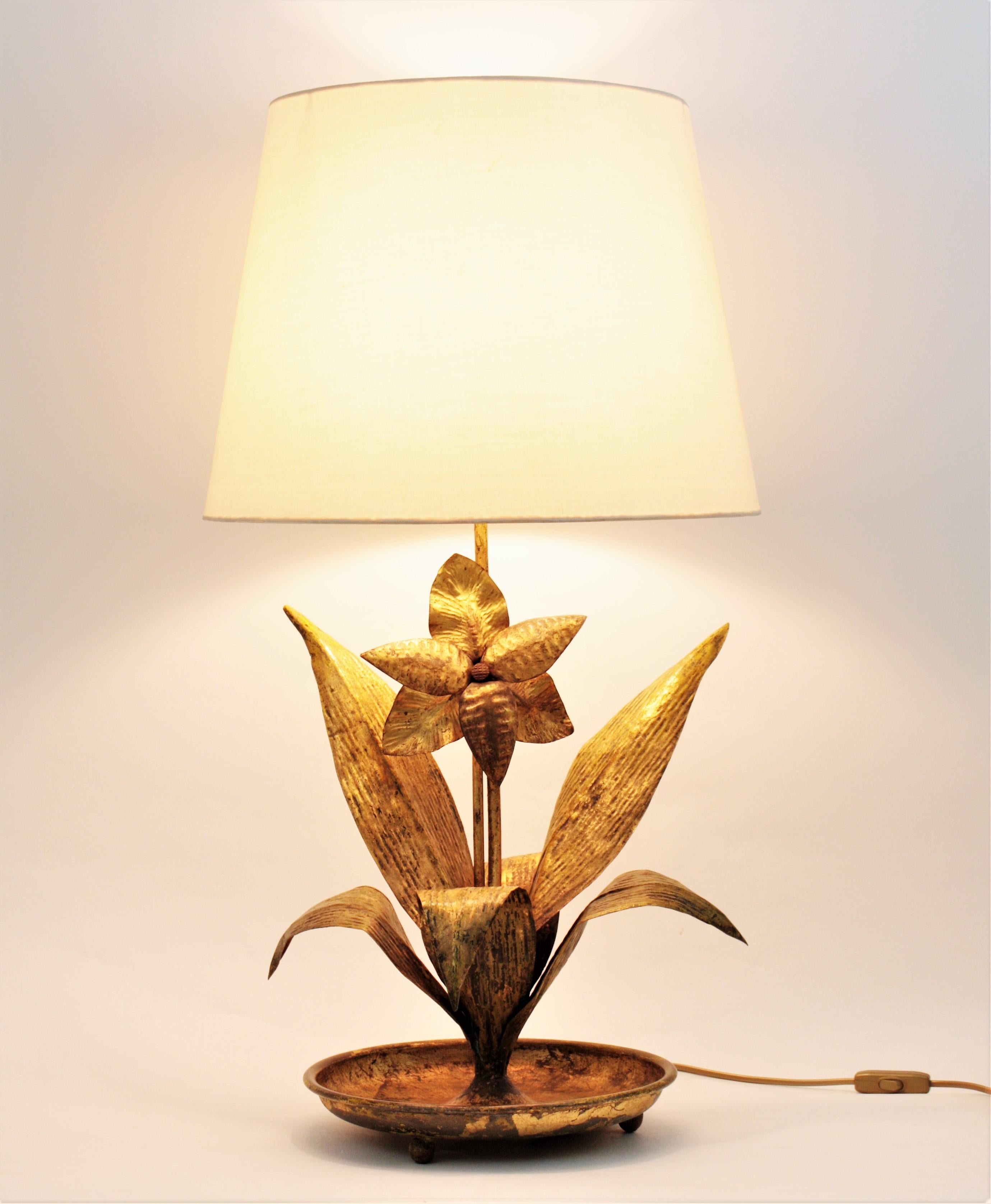 Handcrafted gold leaf gilt metal plant shaped table lamp. France, 1940s.
This elegant table lamp features a plant with large leaves and a central flower detail. The plant rests on a round shaped base with three small ball shaped feet. This lamp has