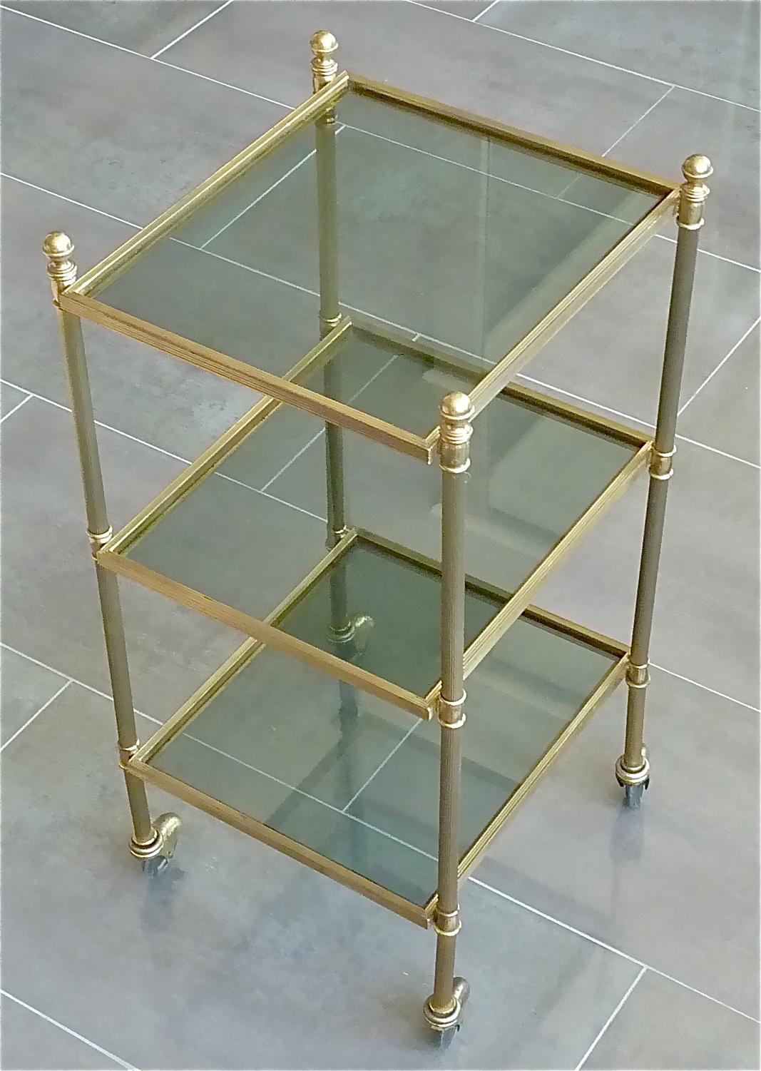 Amazing 3-tiers vintage bar cart, drinks serving trolley or side table on four wheels made by Maison Jansen Paris, France circa 1950. It is made of patinated brass with clear slightly grey to green tinted glass. The small beautiful and useful table