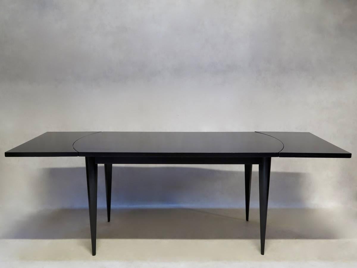 Minimalist dining table, lacquered with a high-gloss black paint finish (with a slightly metallic sheen to it). The table is raised on elegantly slender, tapering legs. The ends of the table are rounded without the extensions, and straight with them