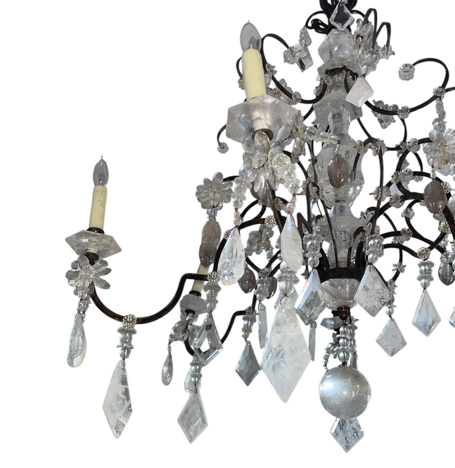 Elegante lampadario francese degli anni '50 realizzato in ferro battuto con una ricca finitura in metallo patinato. I bracci a spirale sono ornati da vetro tagliato, rosette e una serie di prismi di cristallo di rocca, creando un raffinato gioco di