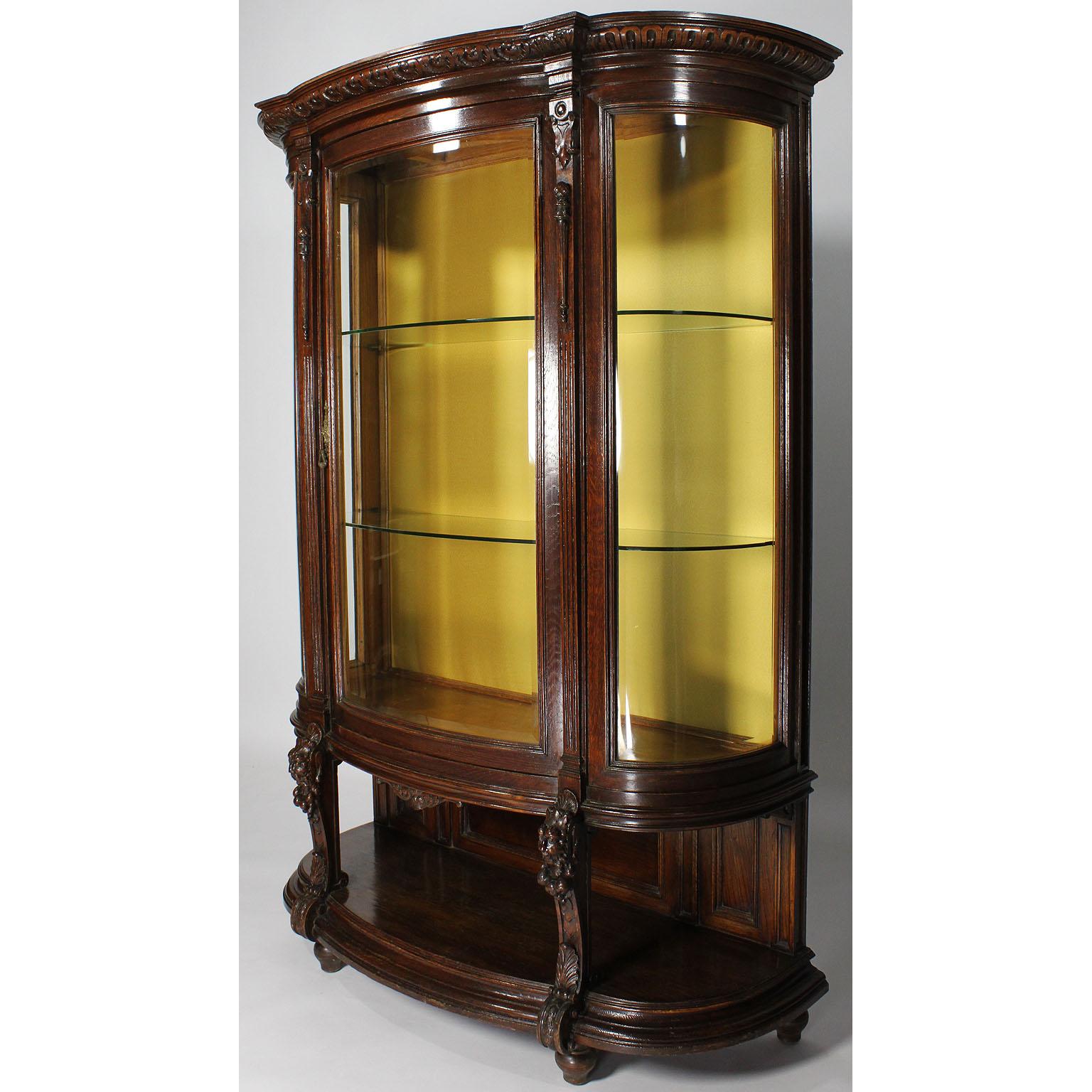 A French 19th-20th century Baroque Revival carved oak Bombé figural vitrine. The single front-door display cabinet with bowed front and side beveled glass panels, with two interior glass shelves and raised on carved front legs with figures of