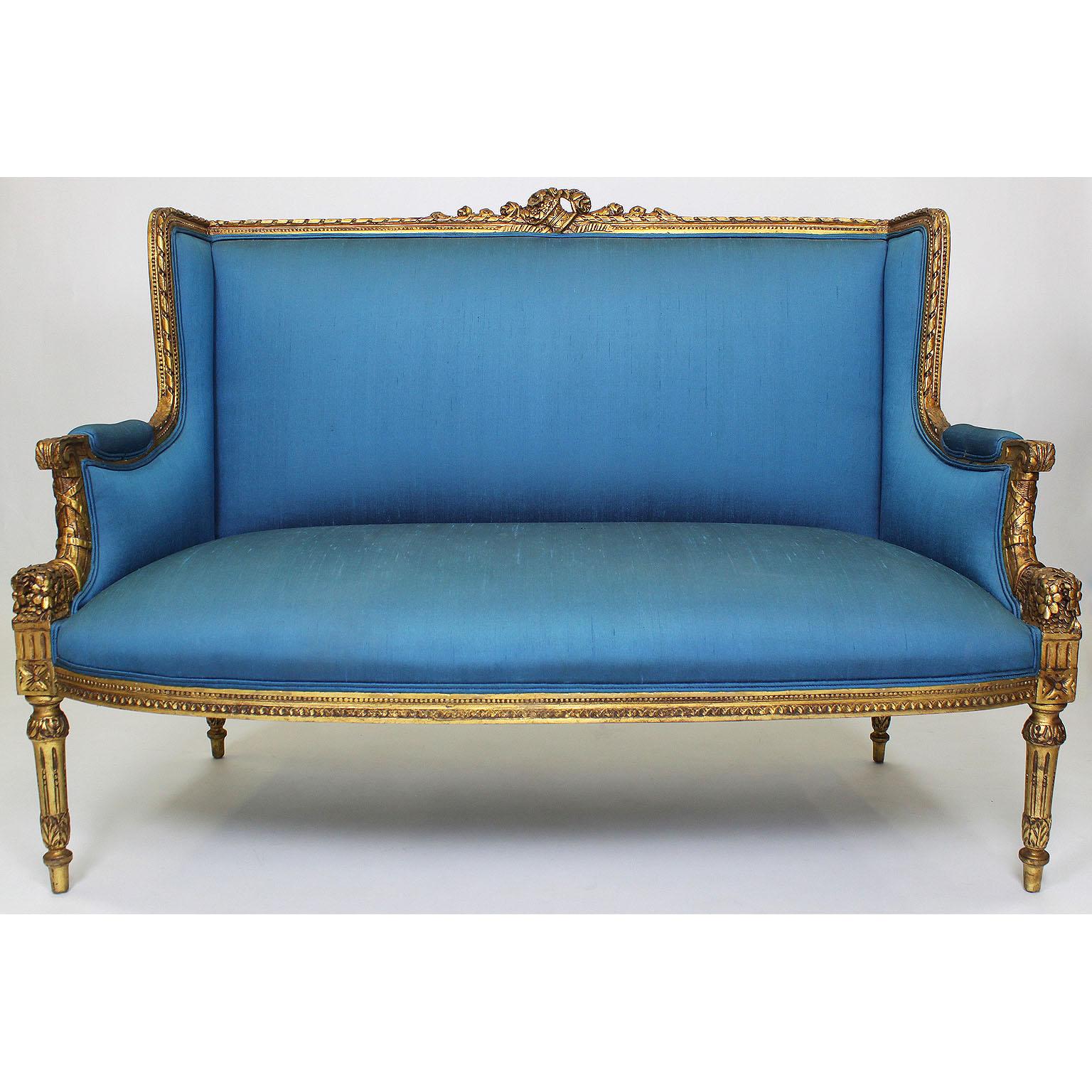 Ensemble de salon en bois doré sculpté de style Louis XVI, comprenant un canapé à deux corps et deux fauteuils, tous garnis de soie bleue de Paris, vers 1900.

La soie présente quelques taches de stries.

Mesures : Hauteur du canapé : 40 3/4 pouces