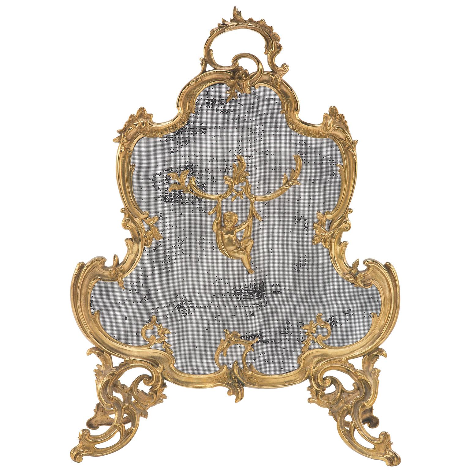 A fine French 19th/20th century Louis XV style gilt-bronze figural fireplace screen. The whimsical ornate ormolu frame with scrolled and rounded corners, detailed with flowers and acanthus and centered with a figure of a Putto suspended from a