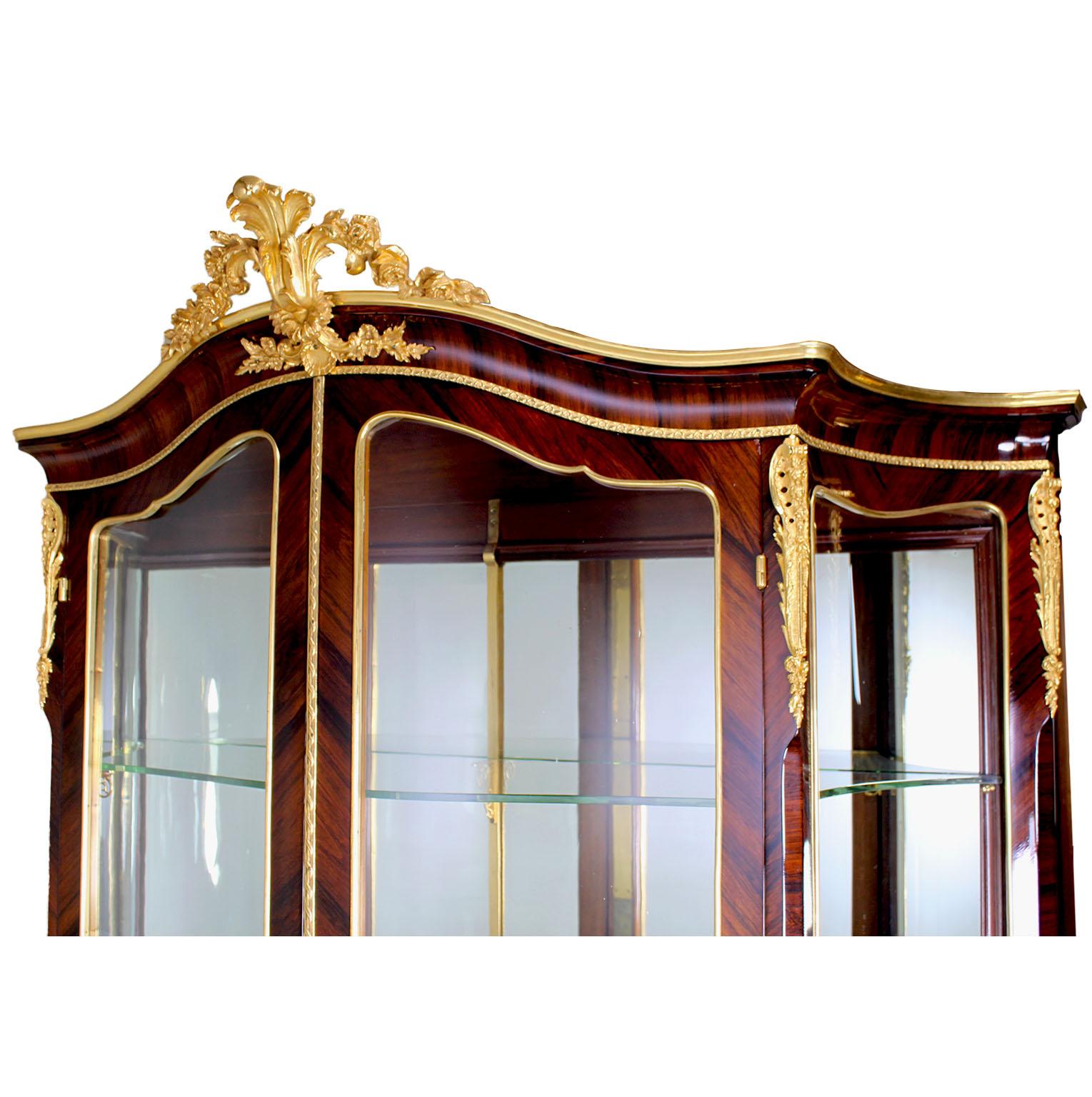 A Fine French 19th-20th century Louis XV style Kingwood and Ormolu mounted two-door vitrine cabinet. The serpentine body with corner ormolu mounts and crowned with an ornate ormolu floral detail centered seashell. The a pair of bent glass front