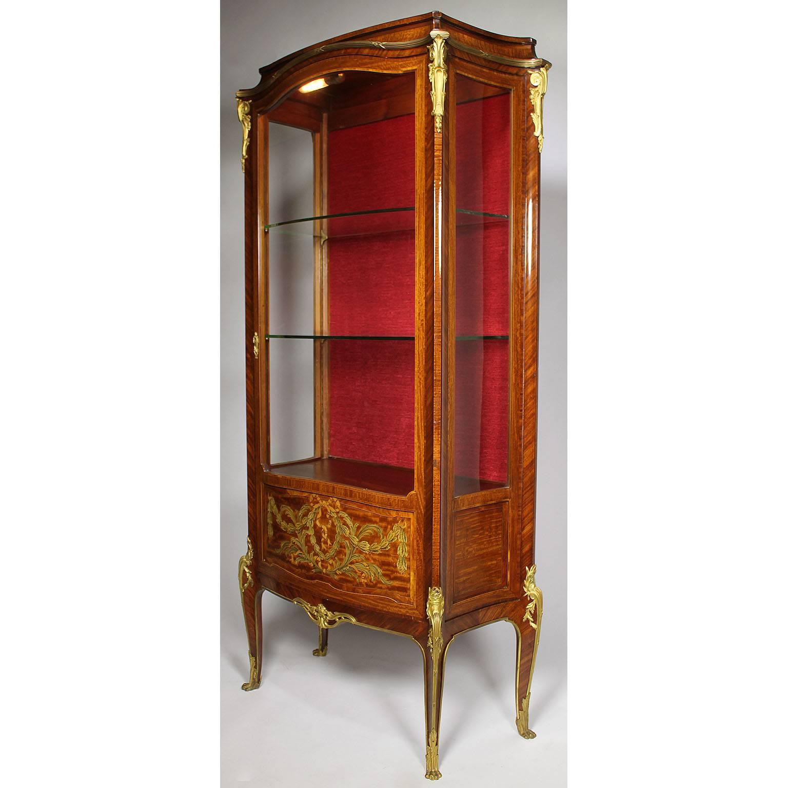 A fine French 19th-20th century kingwood and tulipwood marquetry and gilt-bronze mounted vitrine, in the manner of François Linke (1855-1946). The slender single door display cabinet with a red-velvet backing and bowed glass panels surmounted with