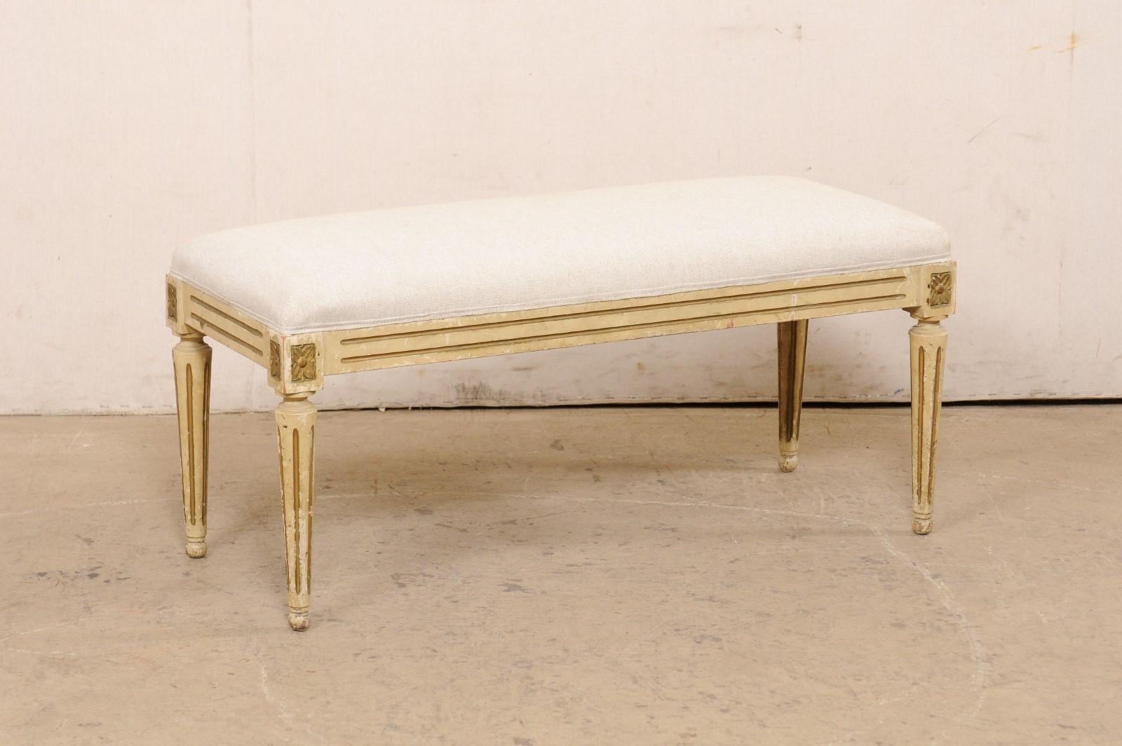 A French Louis XVI style upholstered bench, with its original painted finish, from the 19th century. This antique bench from France is designed in the Louis XVI style with flute carvings about the skirt, rosettes carved at each knee, and presented