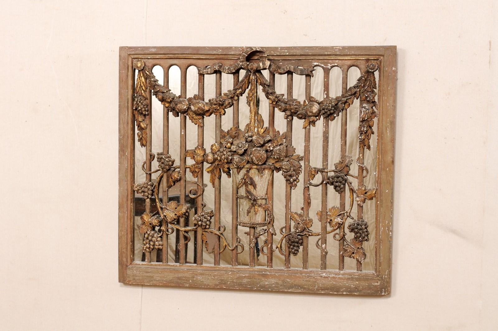 A French carved wood panel, with mirrored back, from the 19th. This antique wall decoration from France features a near-square shaped wooden frame, craved with a floral and vining grape bouquet at center, a foliate swag at top with ribbon, and