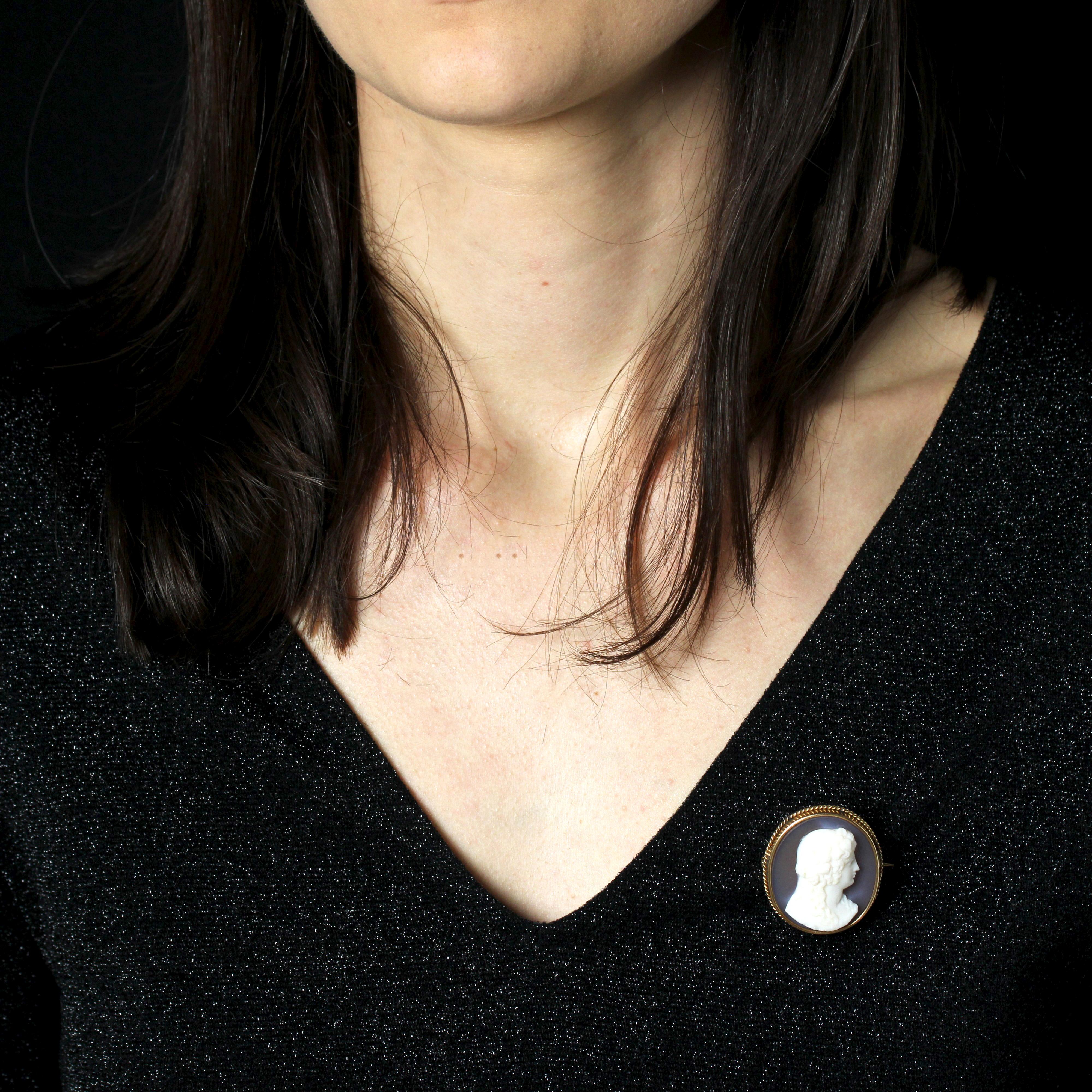 Brooch in 18 karat yellow gold.
This elegant cameo brooch is set in an oval shape in yellow gold and edged with a small gold cord. In the center, a cameo on agate depicts a young woman in profile, with long, loose hair and dressed hairstyle. The