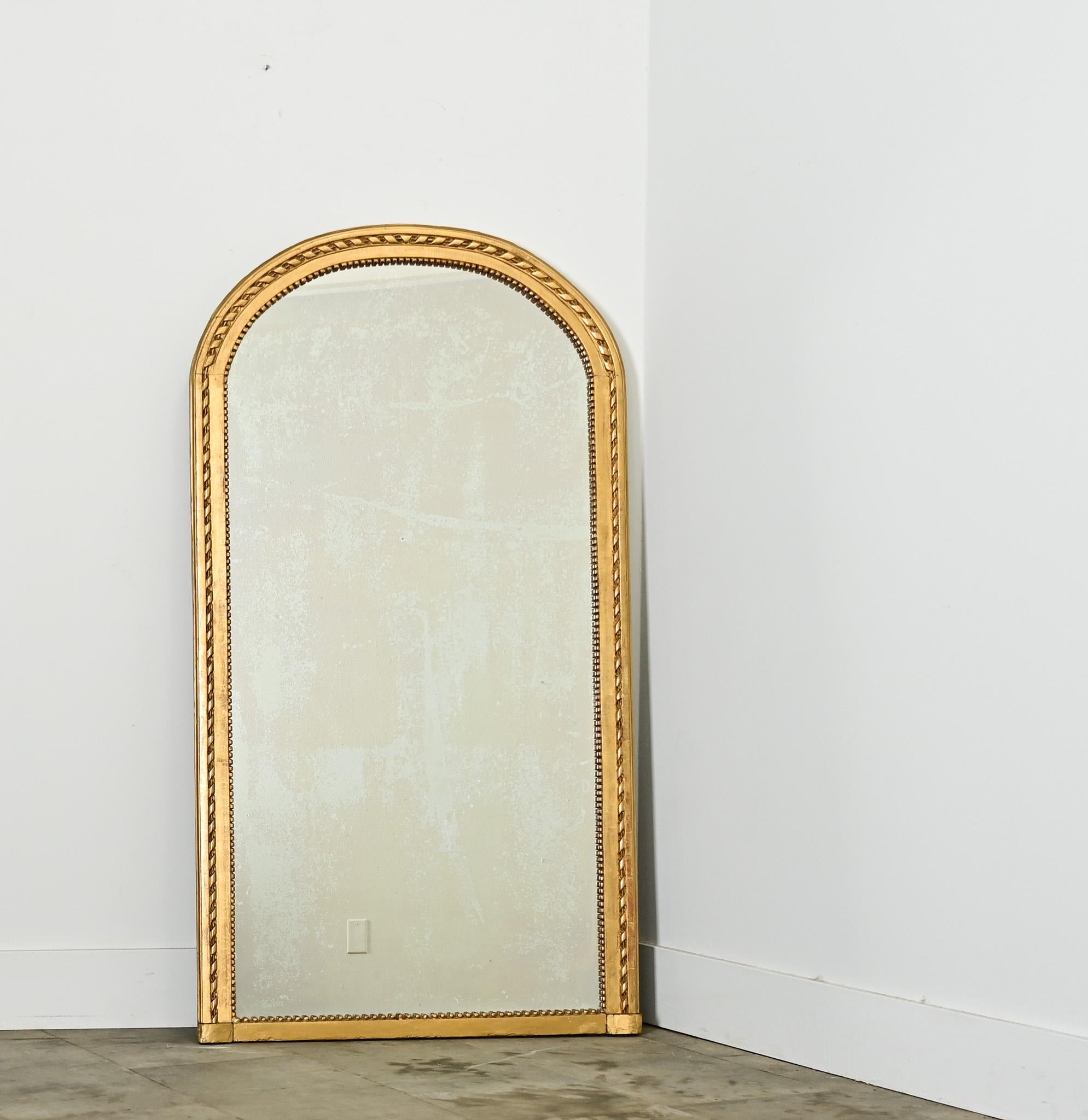 A lovely gilt mantel mirror from 19th century France, the tall proportions of the arch-top frame are emphasized by ribbon-twist and dentil molding around the original mirror plate. Foxing of the glass appears in a translucent haze without