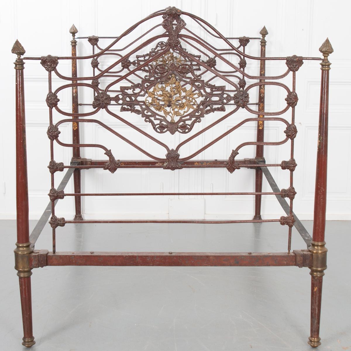 Brass finials adorn the four posts of this vintage French iron and brass bed. The finials are quite fragile as one is broken. The metal bed has remnants of red paint with accents of brass. The center of the headboard and footboard have brass floral