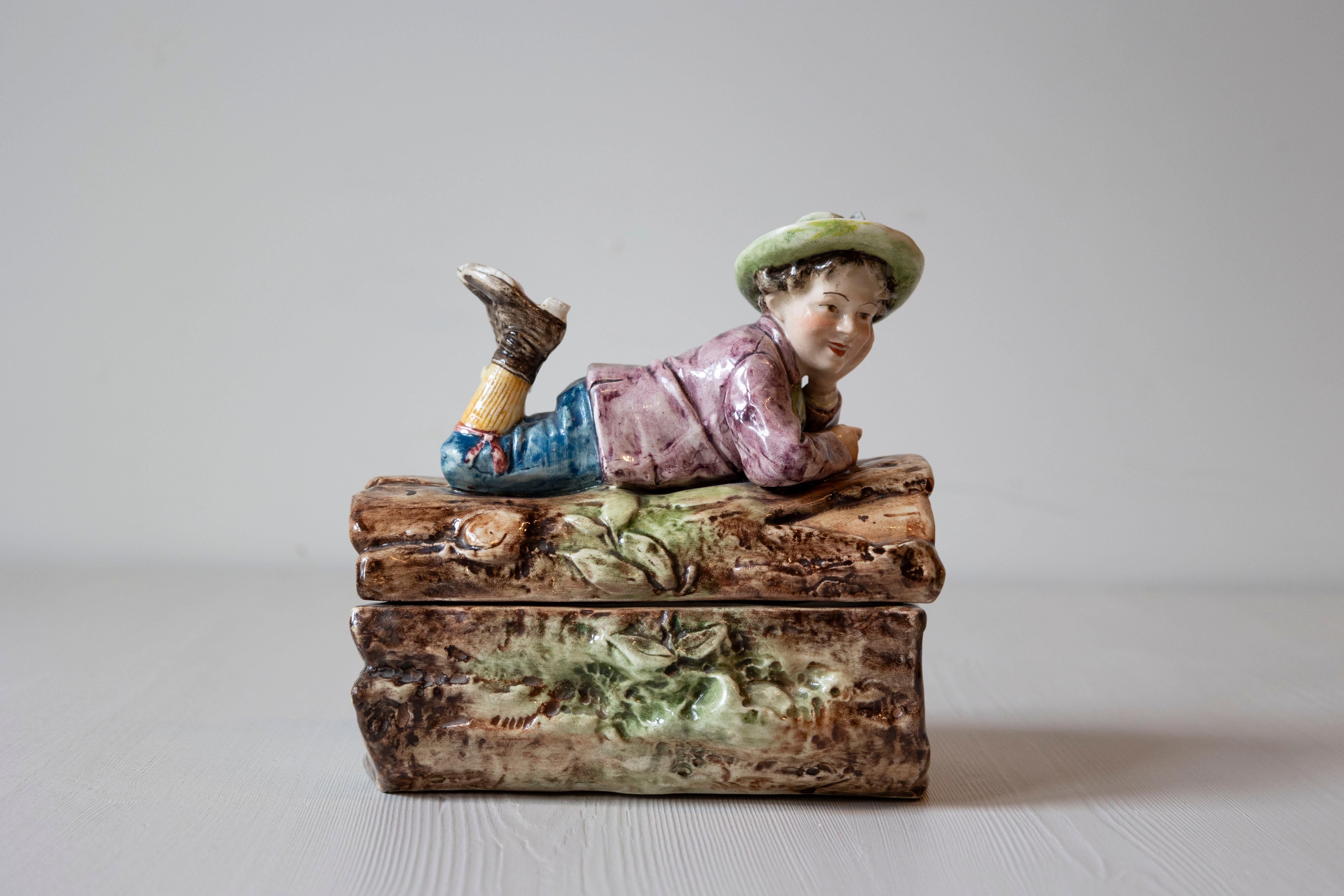 A French barbotine bonbon box from the 19th century, depicting a young boy laying on a tree trunk. Born in France during the 19th century, this charming candy box features a delicate barbotine décor depicting a heartwarming scene: a young boy,