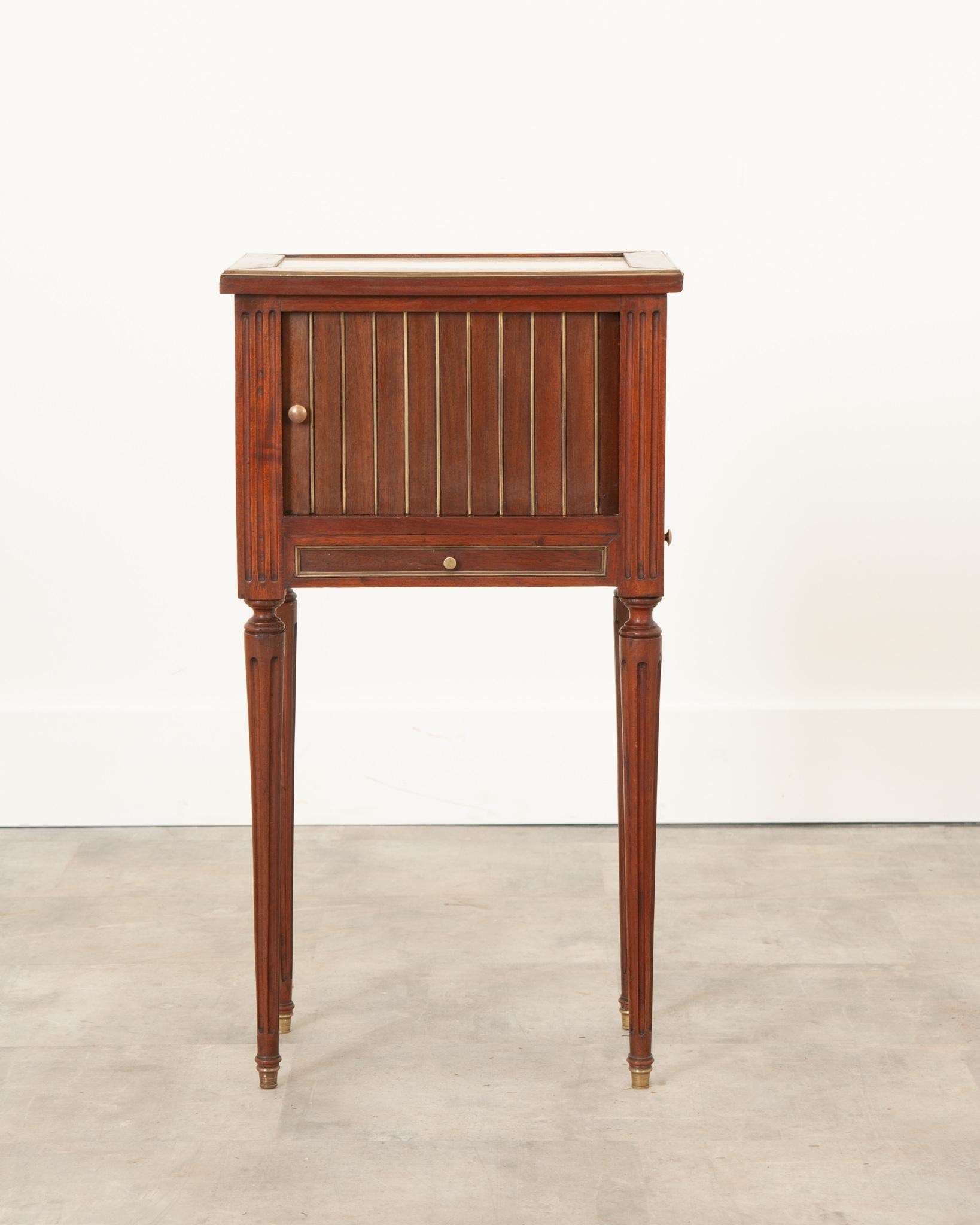 This fantastic little mahogany bedside table was made in 19th Century France, in the Louis XVI style. A white marble top is framed in mahogany which has been trimmed in brass. Brass trim can be found around all panels and drawers. The main cavity is