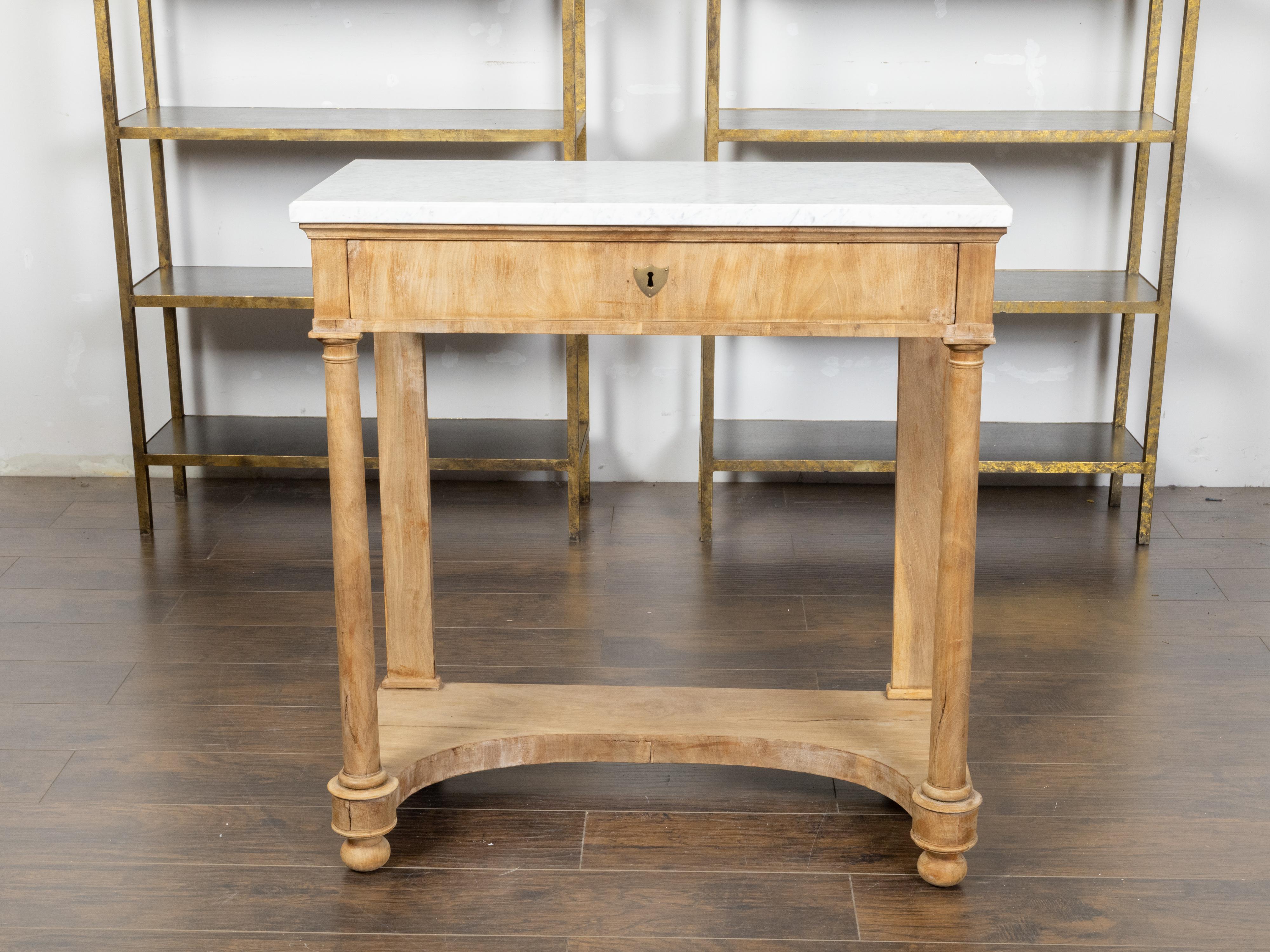 A French bleached walnut console table from the 19th century with white marble top, frieze drawer, in-curving shelf and Doric style column legs. Created in France during the 19th century, this bleached walnut console table features a rectangular