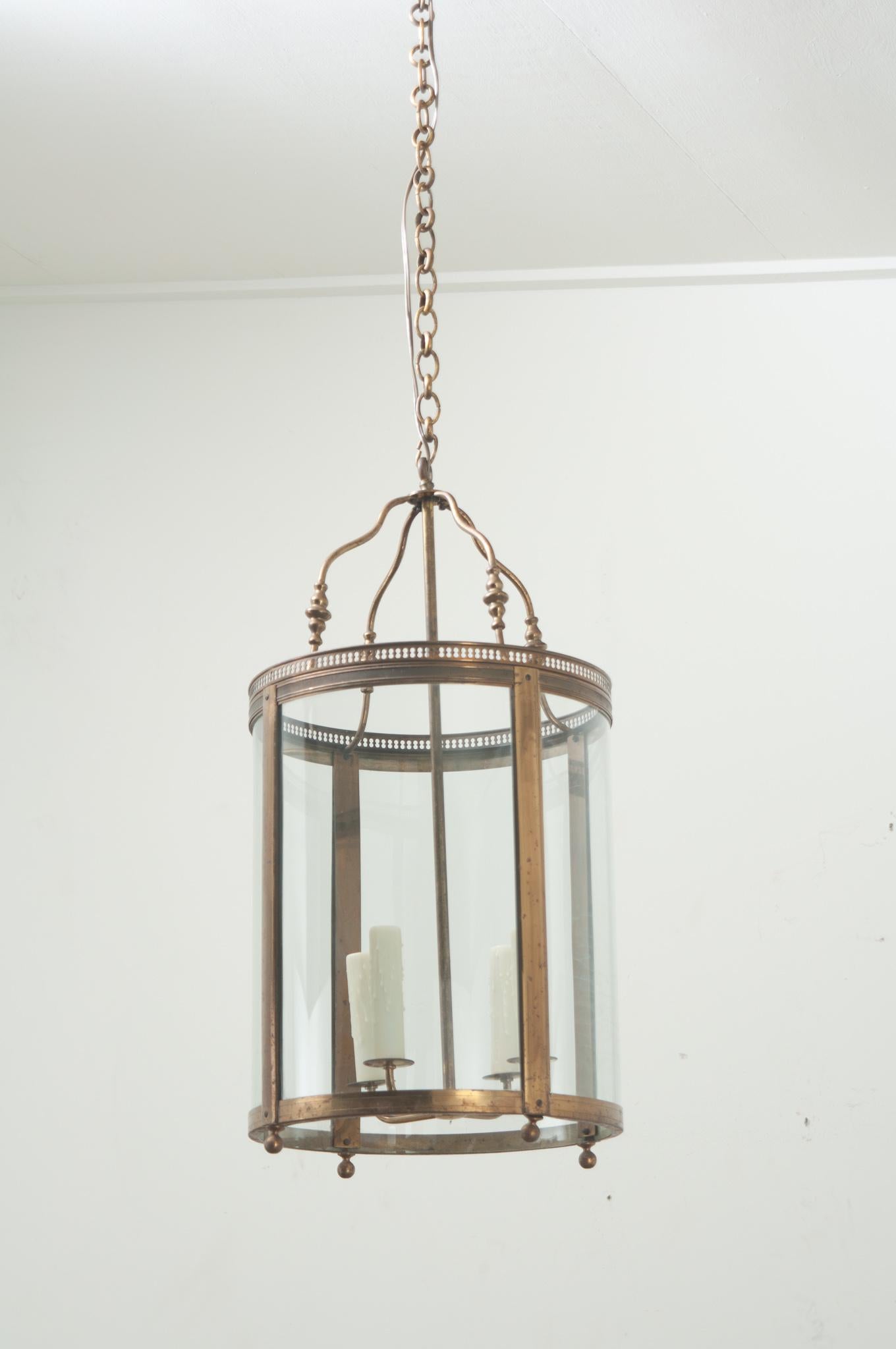 A large French lantern made of brass with curved glass panels. Four detailed arms connect to the chain over a pierced gallery and hold a cluster of four faux candle covers. Cleaned and wired for US electrical using UL listed parts this lantern is