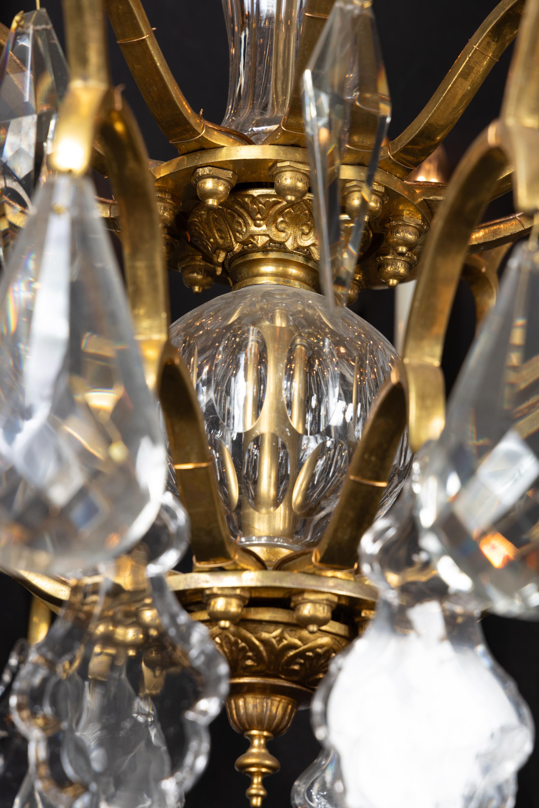 This fine Louis XVI chandelier features beautiful bronze and an impressive crystal center. The 19th century piece sports patterned candle cups, each adorned with crystal plaquettes. Note the detailed bronze finial with pattern-work mirrored at the