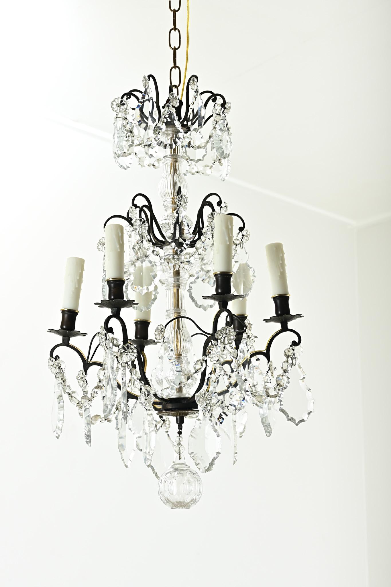 A petite French 6-light bronze and crystal chandelier from the 1800’s. This statement chandelier has a bronze frame with cut crystal drops and cut crystal floral designs hanging from six scalloped chandelier arms with faux wax candle covers. Cleaned
